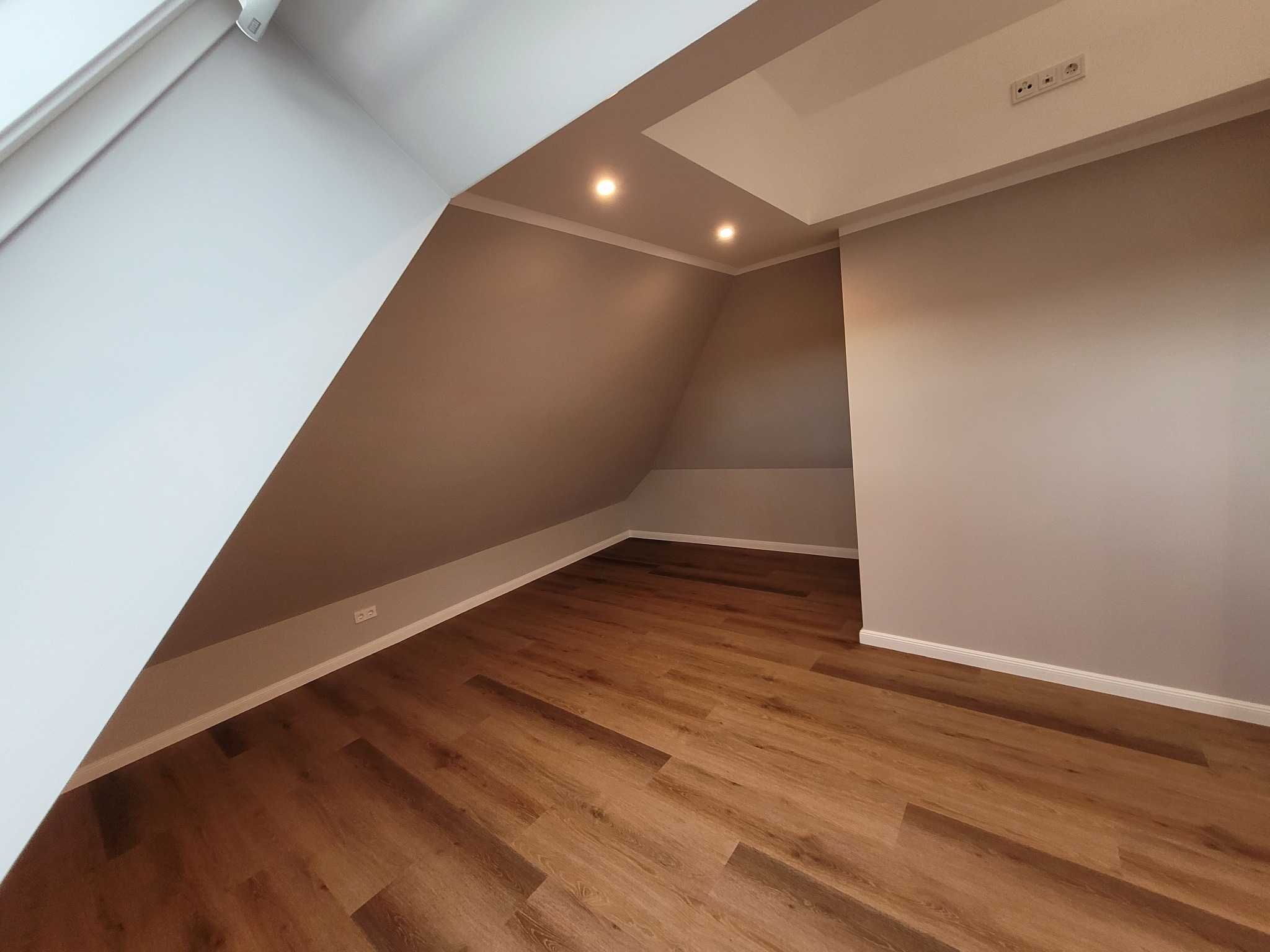 Poddasze po remoncie: skośna ściana z oknem dachowym, drewniana podłoga, szare ściany z białymi listwami przypodłogowymi i sufitowymi, oświetlenie punktowe w suficie.