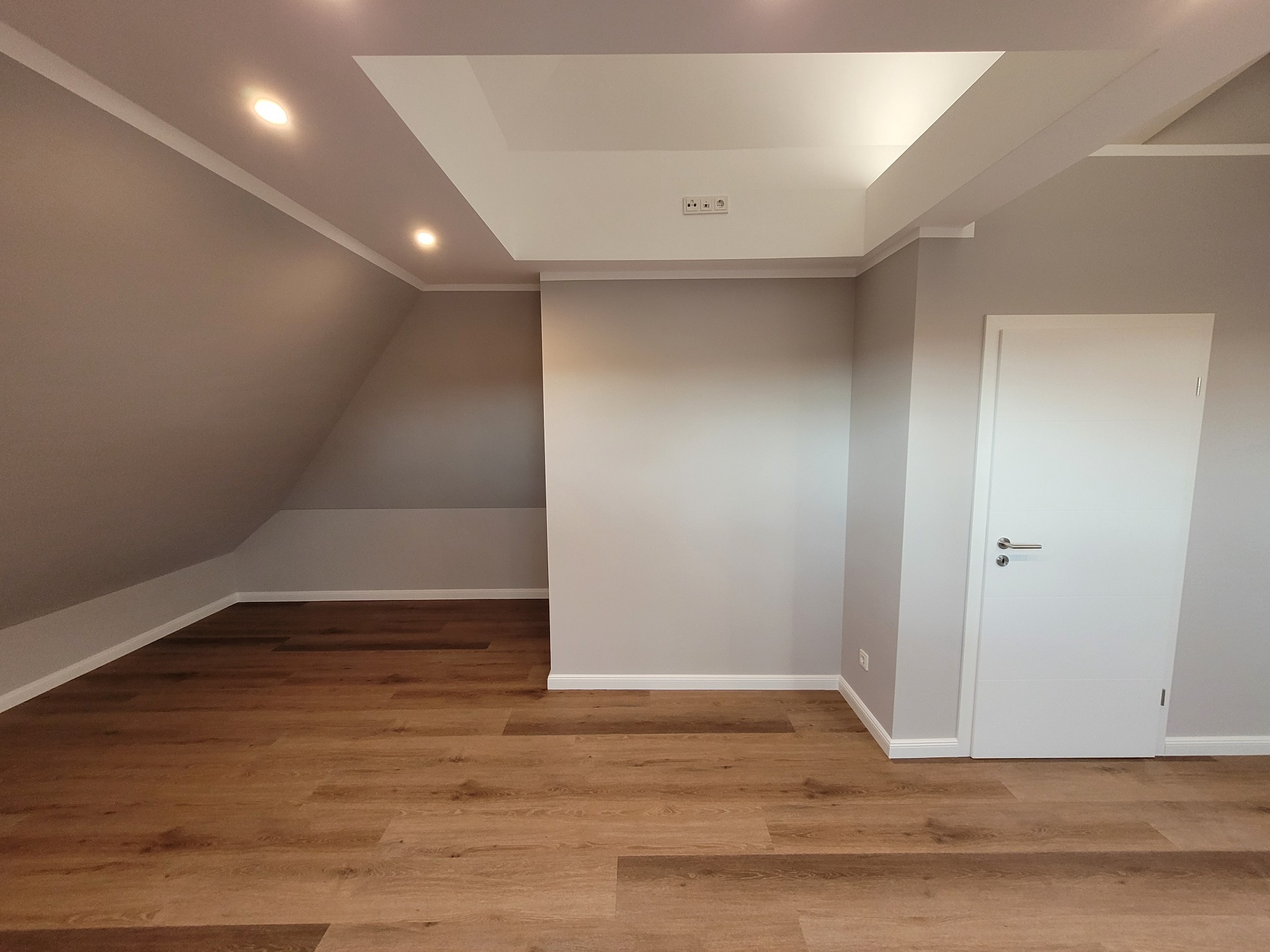 Poddasze po remoncie: jasna drewniana podłoga, szare ściany z białymi listwami przypodłogowymi, białe drzwi z klamką, oświetlenie punktowe w suficie podwieszanym.