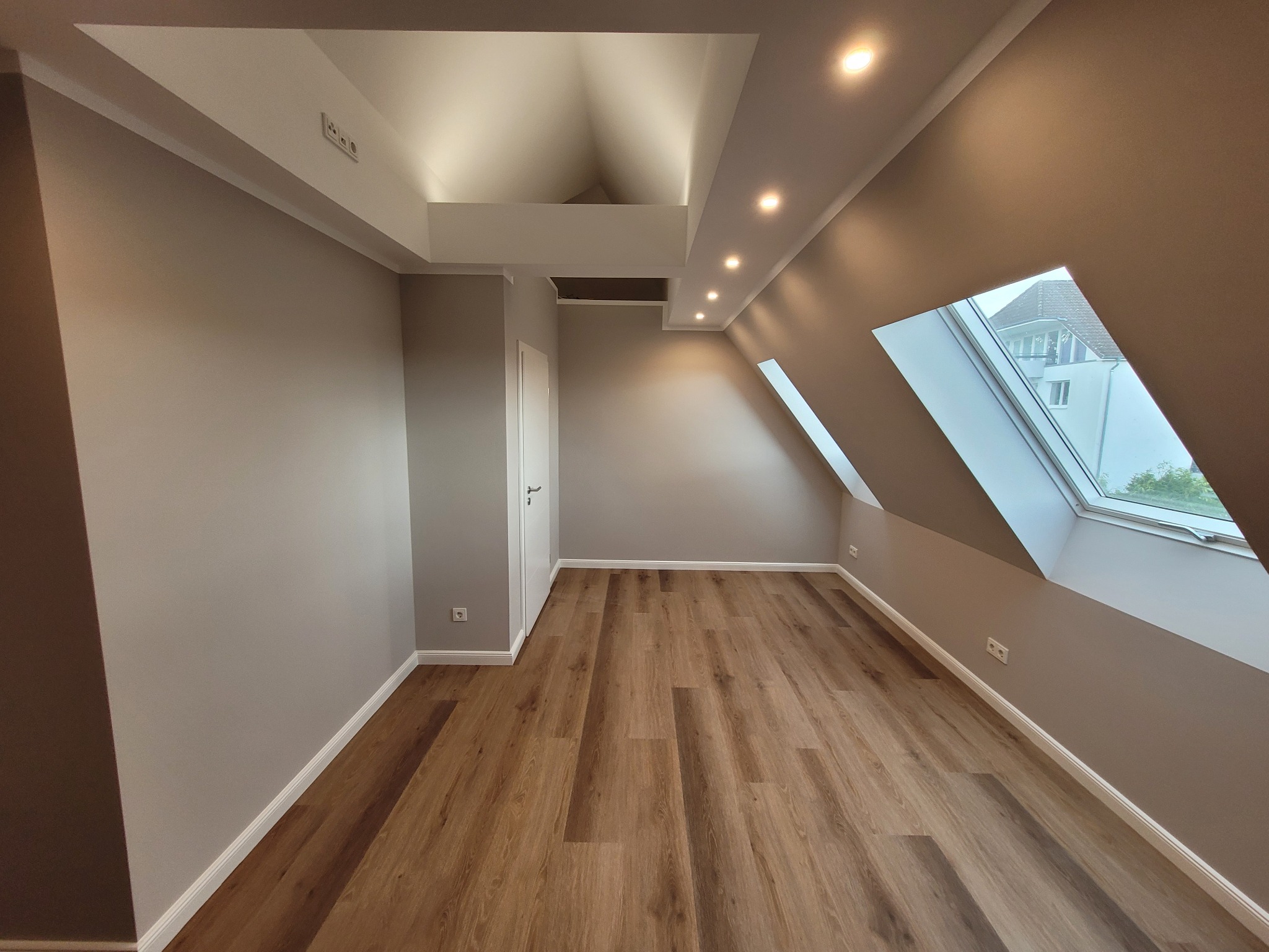 Poddasze po remoncie: Ściany pomalowane na szaro, drewniana podłoga, okno dachowe, białe listwy przypodłogowe i sufit z oświetleniem punktowym.