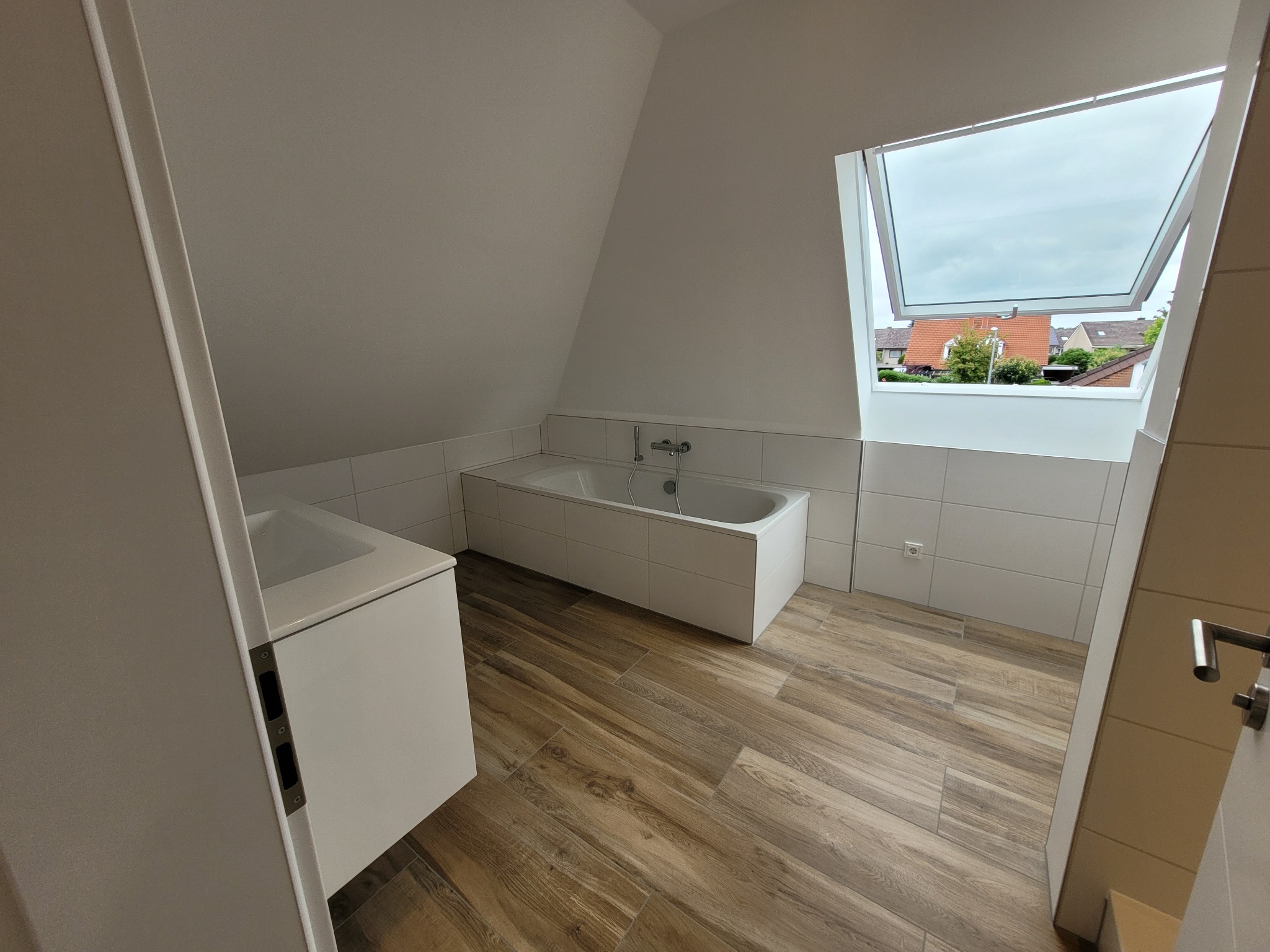 Nowoczesna łazienka na poddaszu z drewnopodobną podłogą, białą zabudową wanny i umywalki oraz oknem dachowym z widokiem na okoliczne domy.