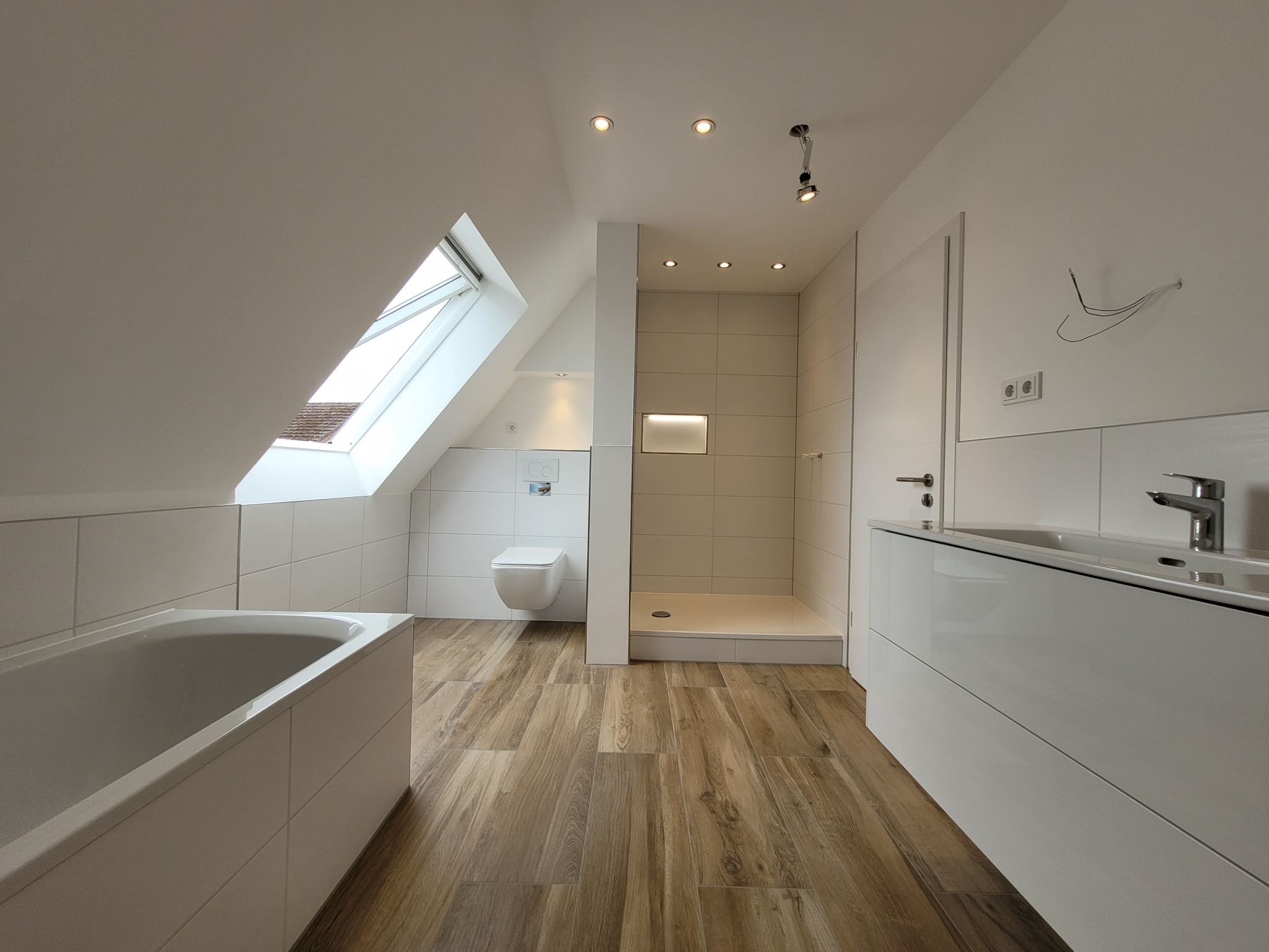 Jasna łazienka na poddaszu z drewnopodobną podłogą, białą wanną, toaletą pod oknem dachowym, kabiną prysznicową i szafką z umywalką.