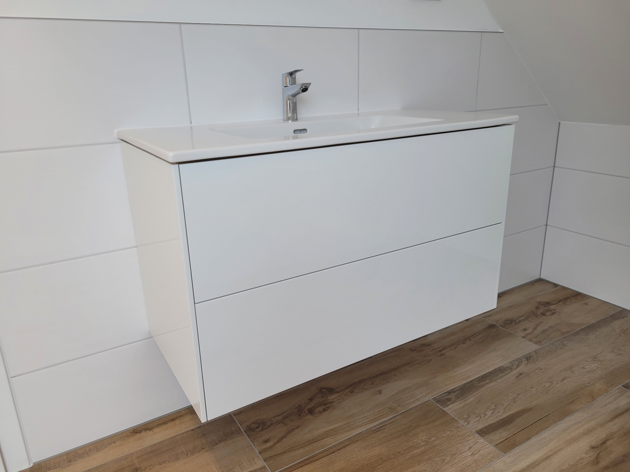 Biała szafka pod umywalkę z chromowaną baterią na tle białych płytek ściennych i podłogi imitującej drewno w łazience na poddaszu.