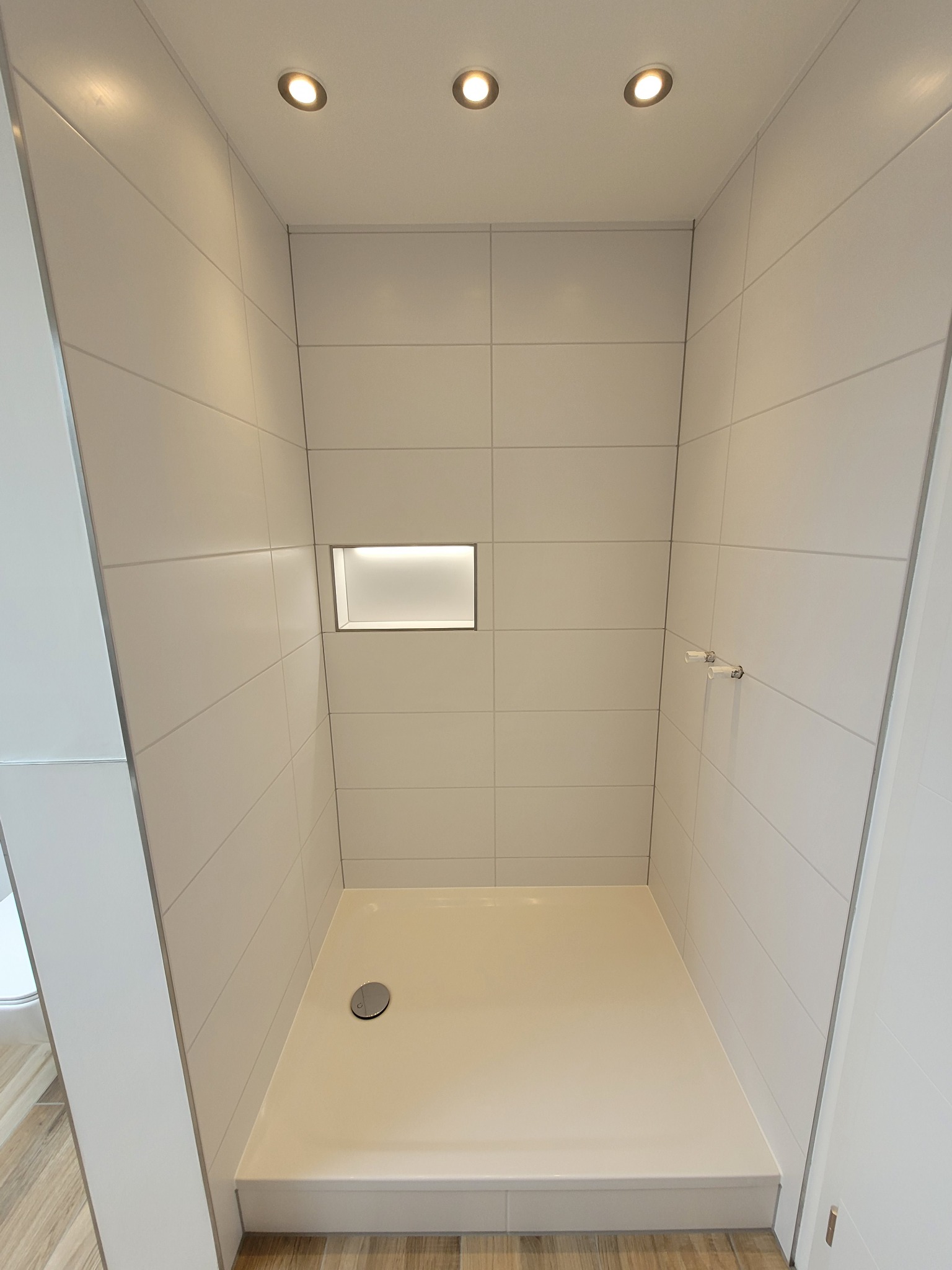 Nowoczesna kabina prysznicowa wyłożona białymi, poziomymi płytkami, z wnęką z podświetleniem, zamontowanymi uchwytami na ręczniki i trzema punktowymi lampami sufitowymi.