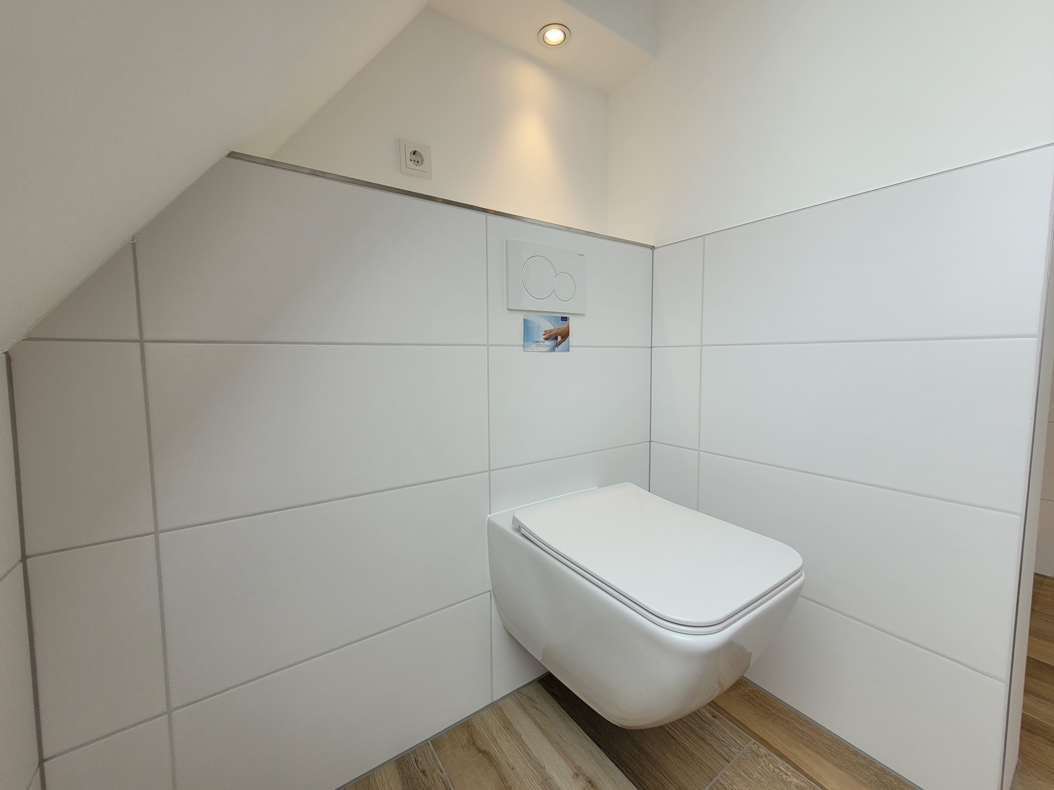 Kompaktowa toaleta podwieszana w nowoczesnej, jasnej łazience na poddaszu, białe płytki ułożone poziomo, drewnopodobna podłoga, minimalistyczny design.