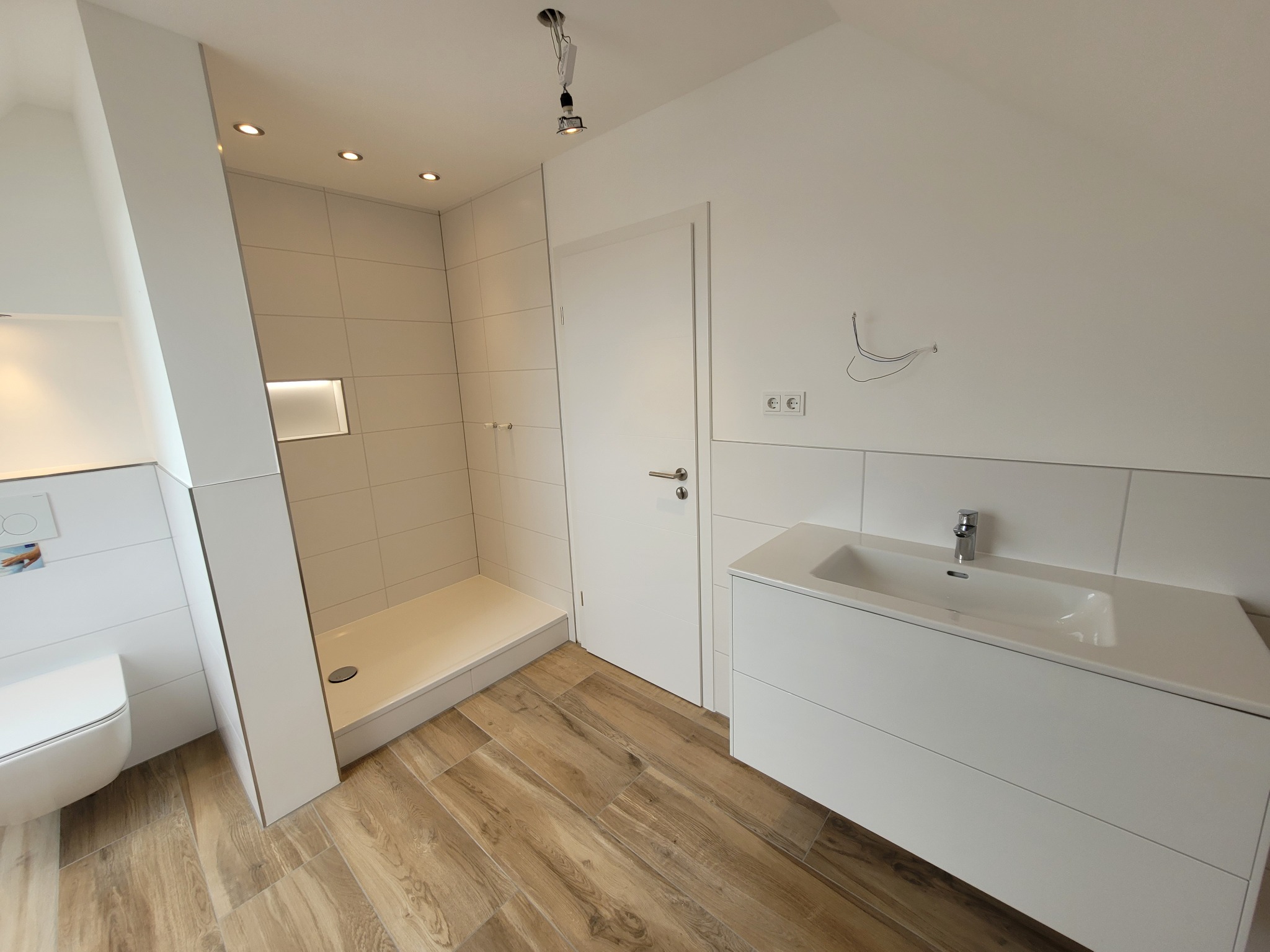 Nowoczesna łazienka z kabiną prysznicową bez brodzika, umywalką z szafką w kolorze białym i podłogą wyłożoną płytkami imitującymi drewno. W tle widoczne niedokończone podłączenie oświetlenia...