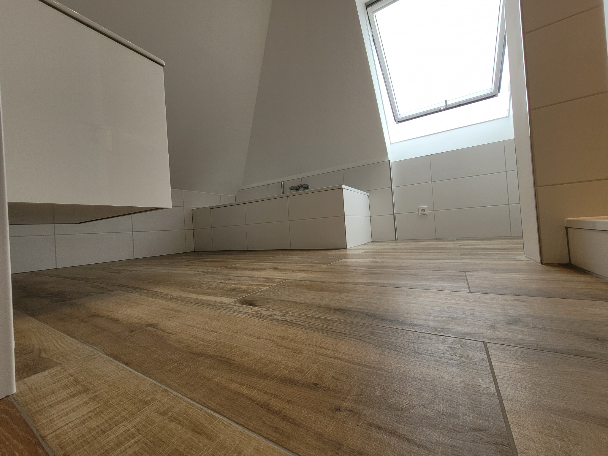 Ukończona łazienka na poddaszu z drewnopodobną podłogą i oknem dachowym, widok z niskiej perspektywy podkreślający przestrzeń i fakturę płytek.