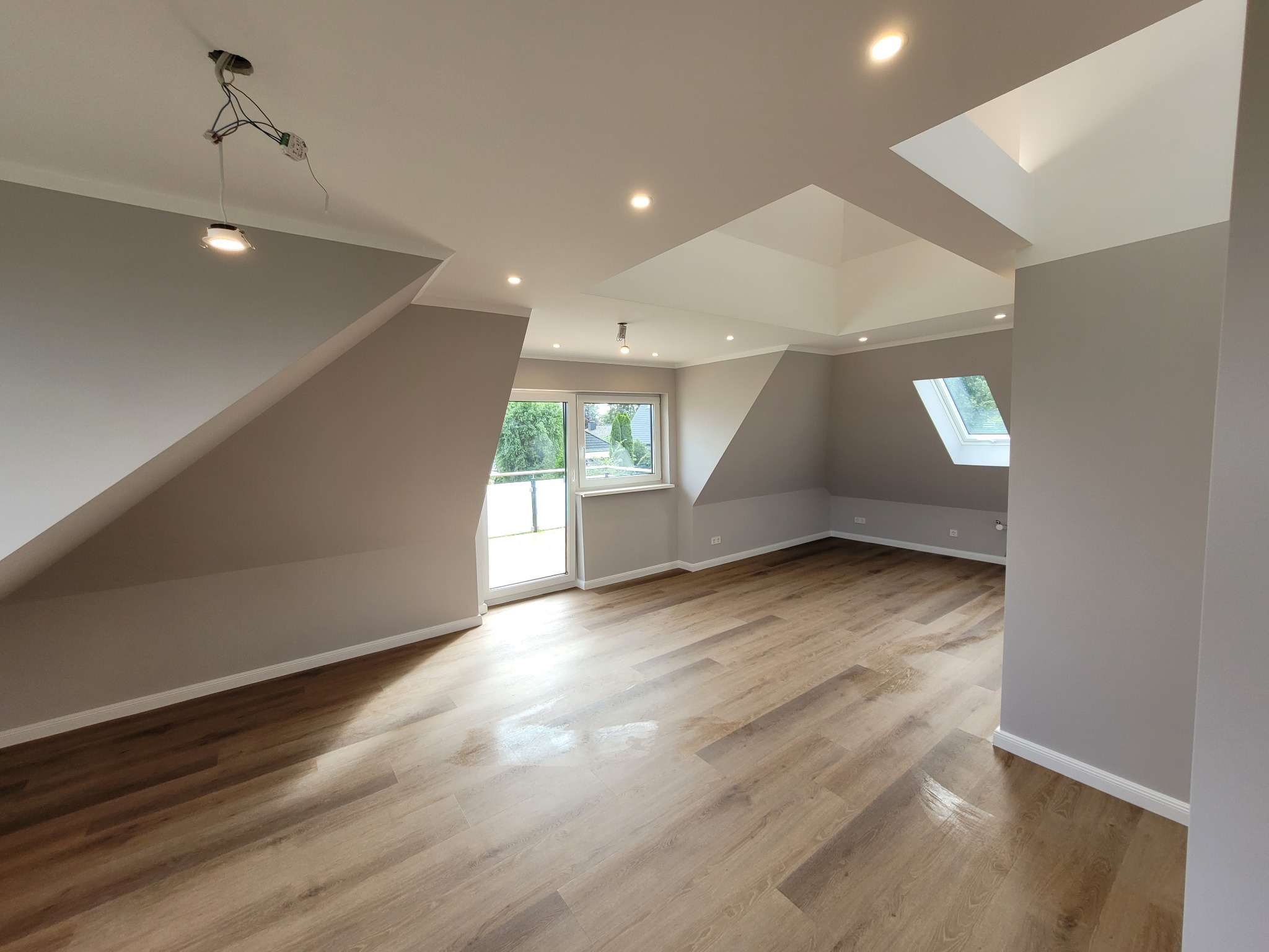 Przestronne, jasne poddasze z drewnianą podłogą, szarymi ścianami i oknami dachowymi, oświetlone punktowo, z widoczną instalacją elektryczną przy jednym z punktów świetlnych.