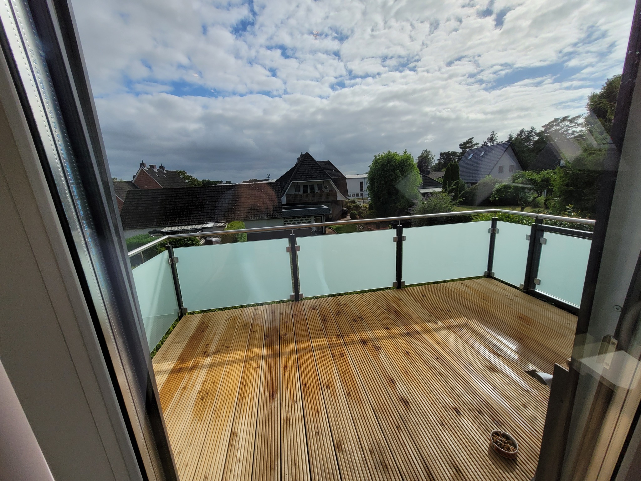 Drewniany taras na balkonie z przeszkloną balustradą, widok na domy i pochmurne niebo, widoczne detale konstrukcyjne okna.