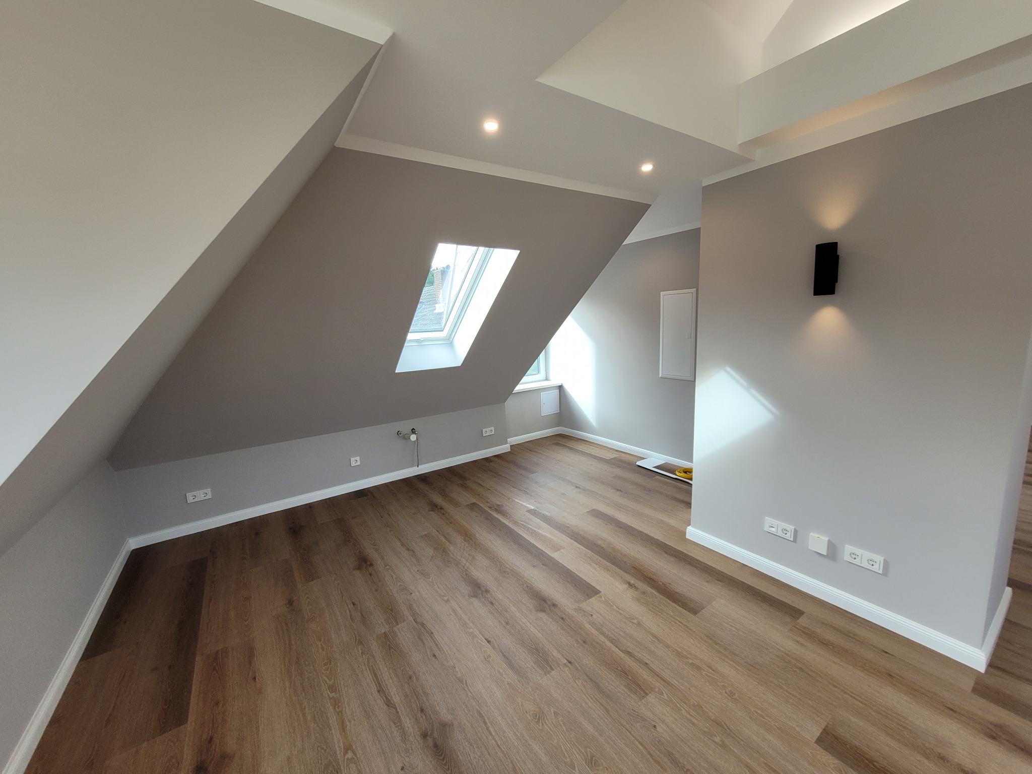 Poddasze po remoncie z drewnianą podłogą, szarymi ścianami i oknem dachowym, oświetlone punktowymi lampami sufitowymi i czarnym kinkietem ściennym.