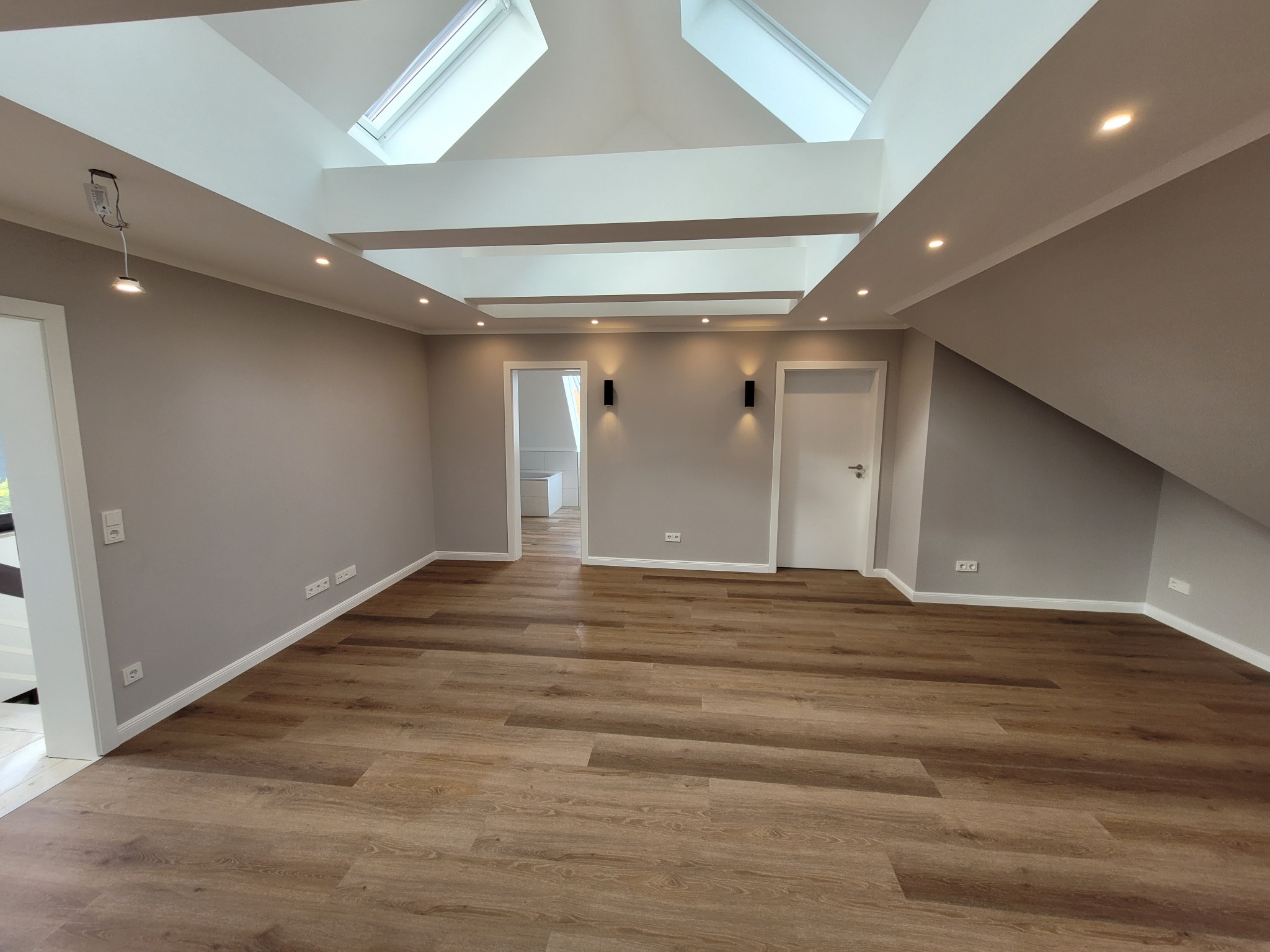 Poddasze po remoncie z drewnopodobną podłogą, szarymi ścianami, białymi drzwiami i oknami dachowymi w suficie, oświetlone punktowymi lampami.