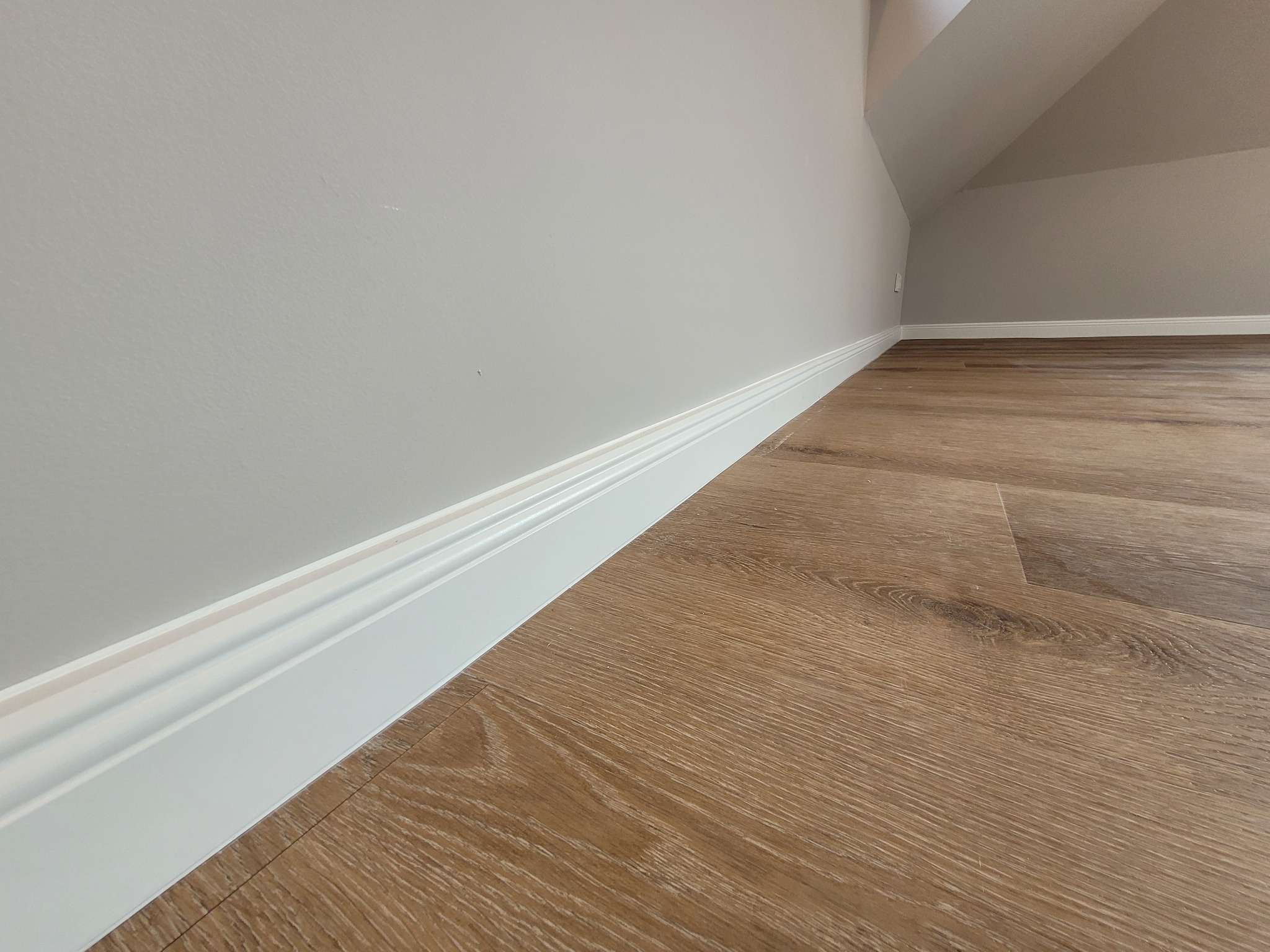 Jasne panele podłogowe imitujące drewno, białe listwy przypodłogowe z frezowaniem, szara ściana pod skosem na poddaszu.