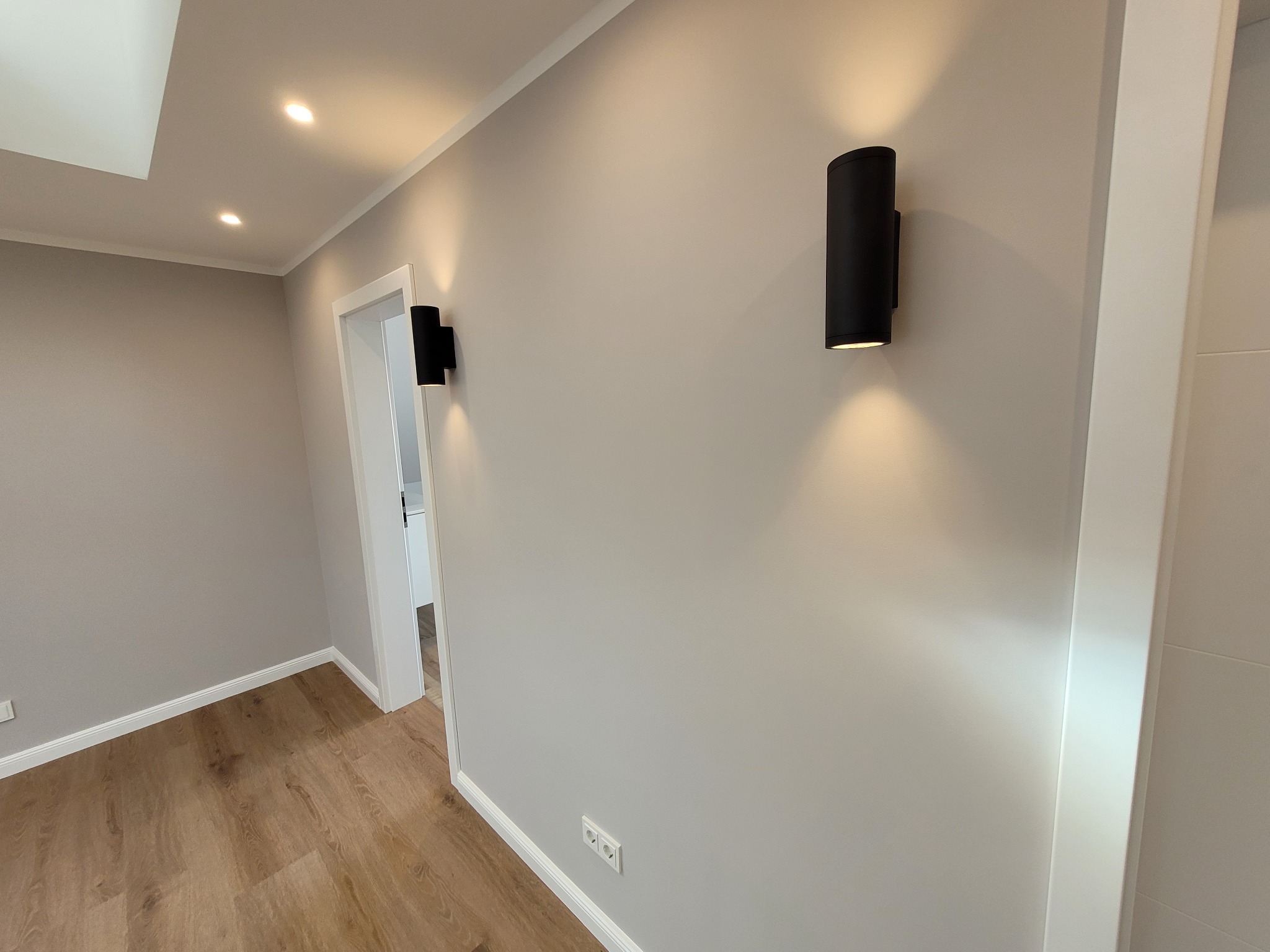 Korytarz w Kotuni z drewnianą podłogą i szarą ścianą oświetloną przez dwa czarne, cylindryczne kinkiety.