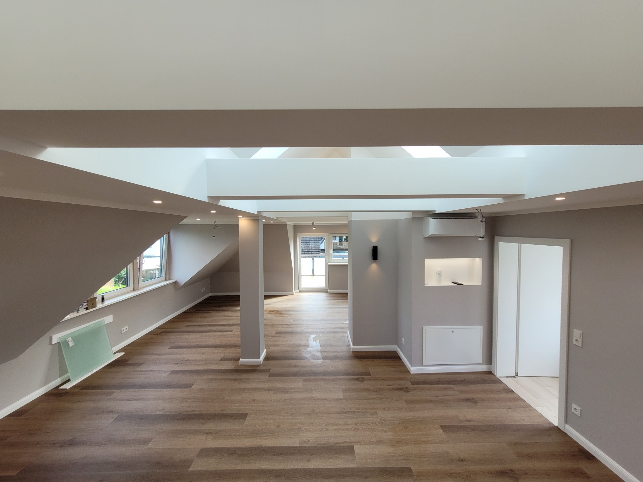 Jasne, przestronne poddasze z drewnianą podłogą, szarymi ścianami i oknami dachowymi, oświetlone punktowymi lampami i nowoczesnym oświetleniem ściennym.