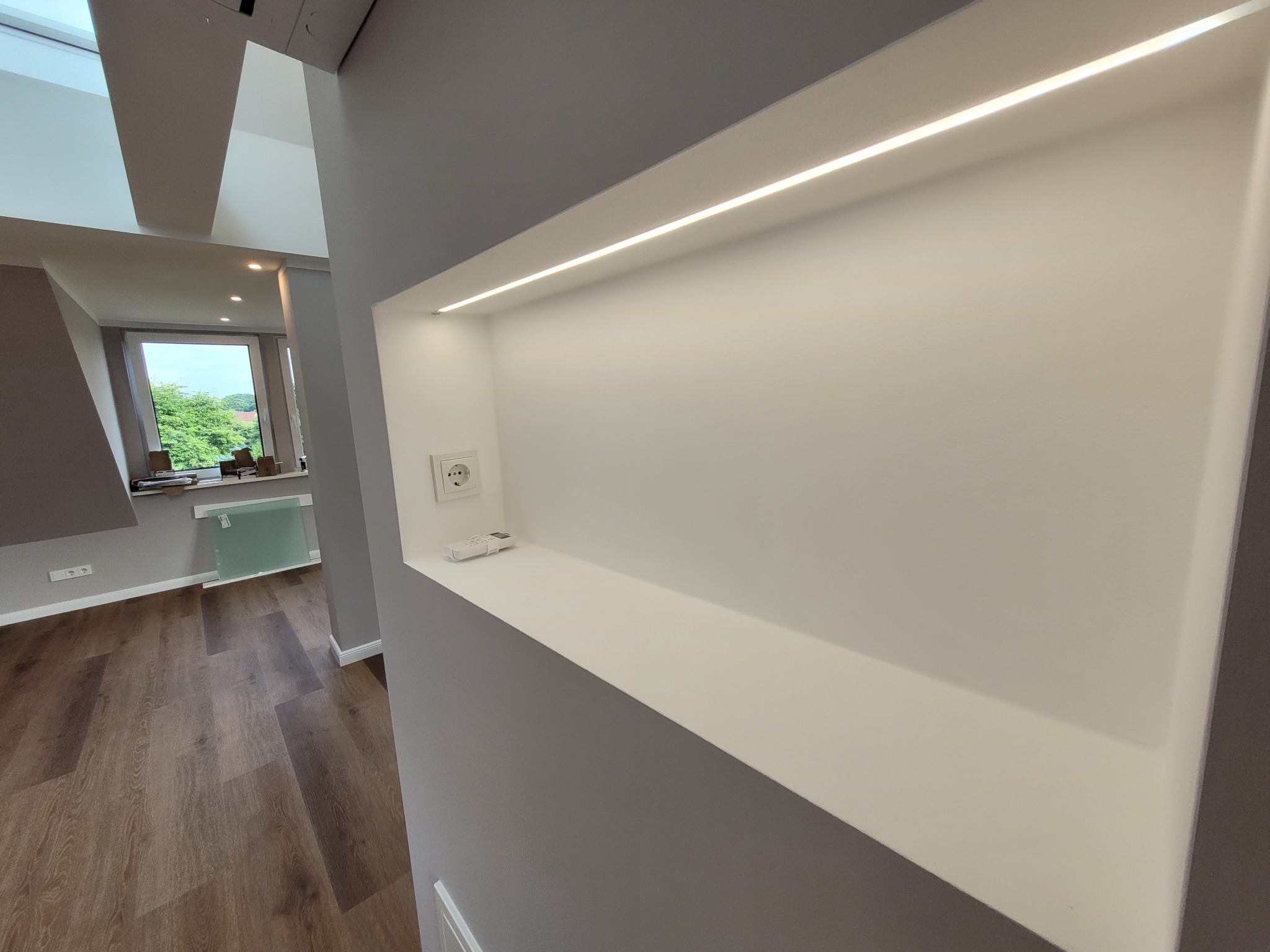 Wnęka w ścianie z oświetleniem LED, gniazdko elektryczne i pilot, widok na pokój z drewnianą podłogą i oknem.