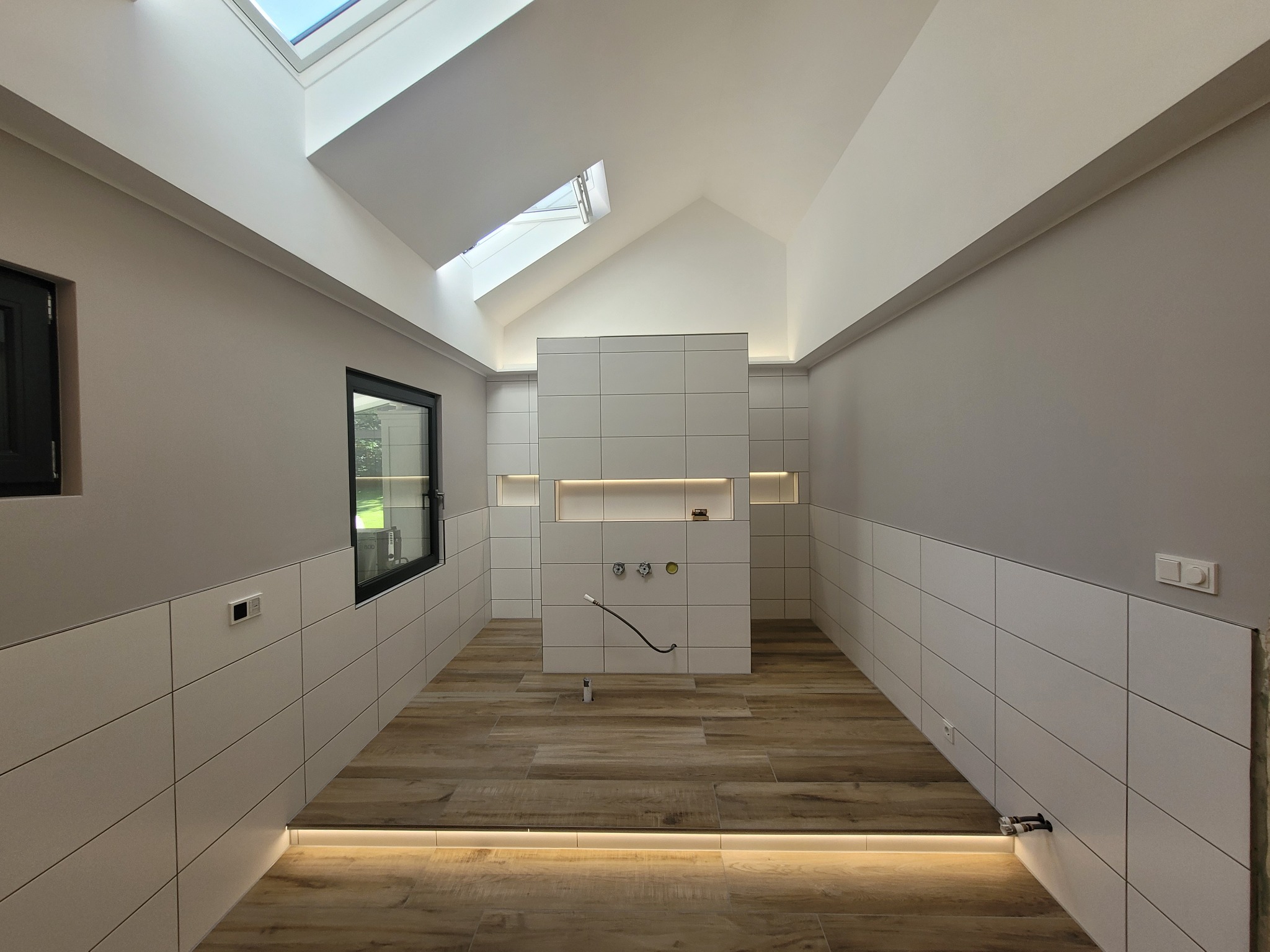 Wnętrze łazienki w trakcie prac wykończeniowych; widoczne białe płytki na ścianach do połowy wysokości, drewnopodobne płytki na podłodze z podświetleniem LED, okno, okna dachowe i przygotowane...