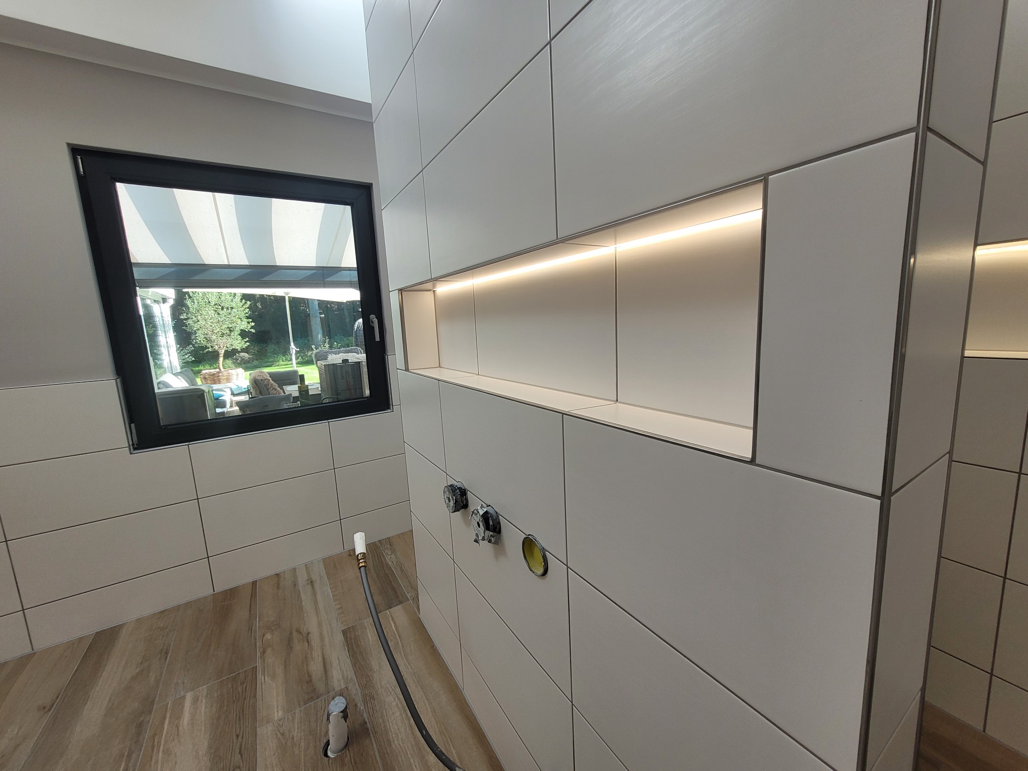 Ściana w trakcie układania płytek w łazience, widoczne przyłącza wodne, wnęka z oświetleniem LED i okno z widokiem na ogród.