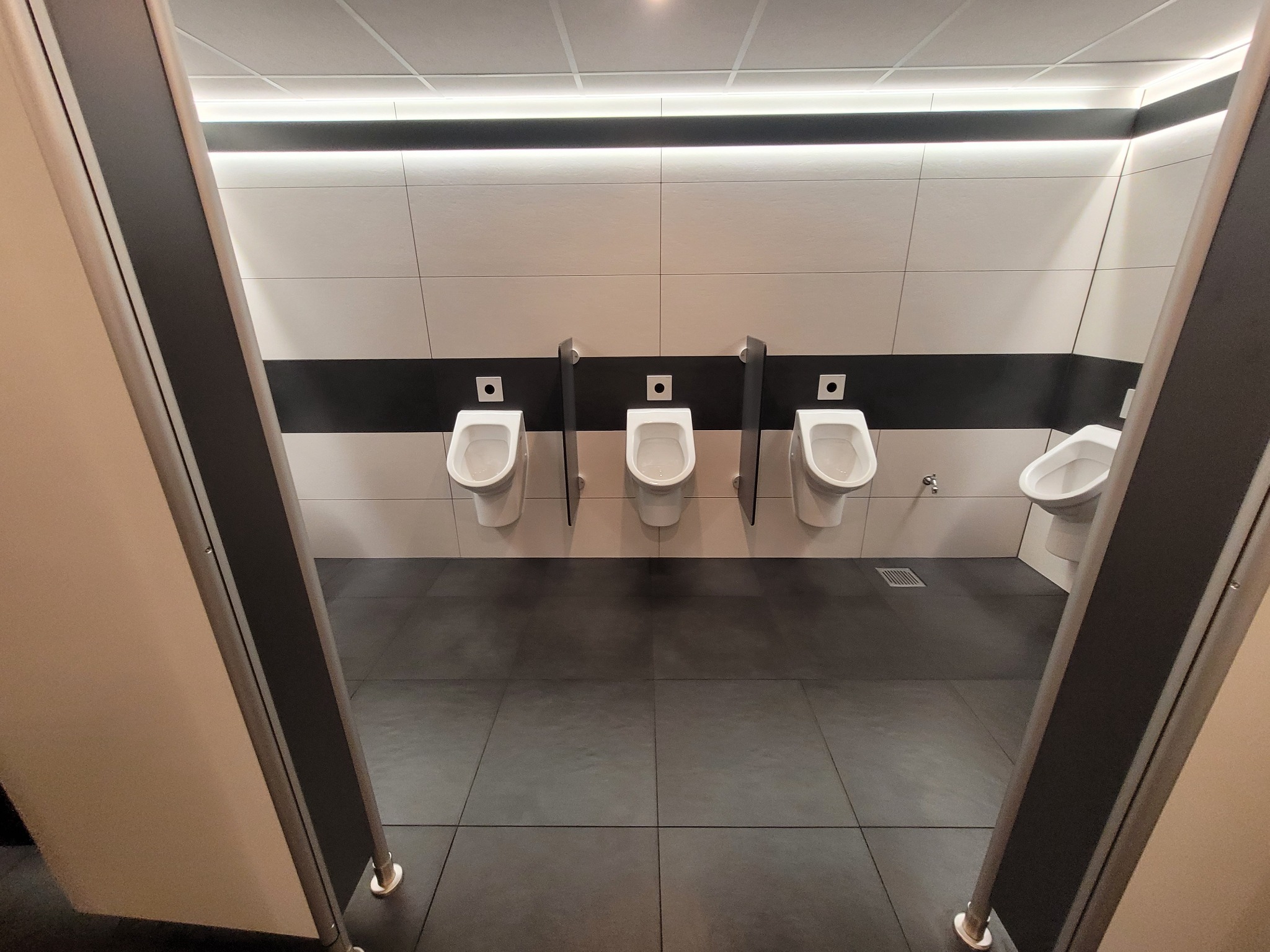 Publiczna toaleta męska z trzema pisuarami na ścianie z białymi i czarnymi płytkami, widok z kabiny, szare płytki na podłodze, oświetlenie LED.