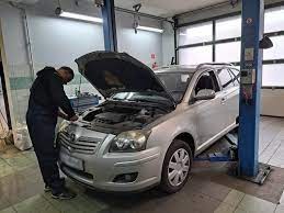 Srebrny samochód kombi na podnośniku w warsztacie, z otwartą maską silnika, przy którym stoi osoba w czarnej bluzie, prawdopodobnie sprawdzająca lub naprawiająca coś w silniku. Widoczne narzędzia...