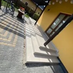 Wykończone schody zewnętrzne z jasnego betonu prowadzące do domu o żółtej elewacji, widok z boku na tle kostki brukowej i roweru.