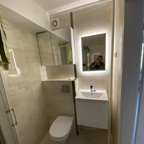 Wykończona łazienka z beżowymi płytkami, podwieszaną toaletą, szafką z umywalką i lustrem z podświetleniem LED, widoczne odbicie osoby robiącej zdjęcie.