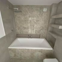 Nowoczesna łazienka z prostokątną białą wanną w zabudowie, szara ściana z efektem betonu, wnęka z półkami i deszczownica