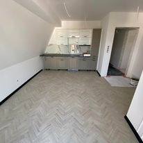 Jasna podłoga z paneli ułożonych w jodełkę francuską w nowym, jeszcze niewykończonym pomieszczeniu ze skośnym sufitem, widoczne przygotowane szafki kuchenne i niedokończone ściany.