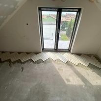 Układanie jasnych paneli w jodełkę na poddaszu, widoczne niedokończone ściany i okno balkonowe z widokiem na zewnątrz.