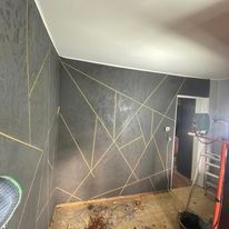 Szara ściana z geometrycznym wzorem wykonanym z cienkich, złotych linii, widoczne ślady po remoncie na podłodze i drabina w tle, oświetlenie mieszane.