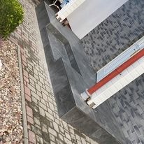 Schody zewnętrzne wyłożone ciemnoszarą płytką, z widocznym fragmentem chodnika z kostki brukowej i elementami elewacji budynku, ujęcie z góry.