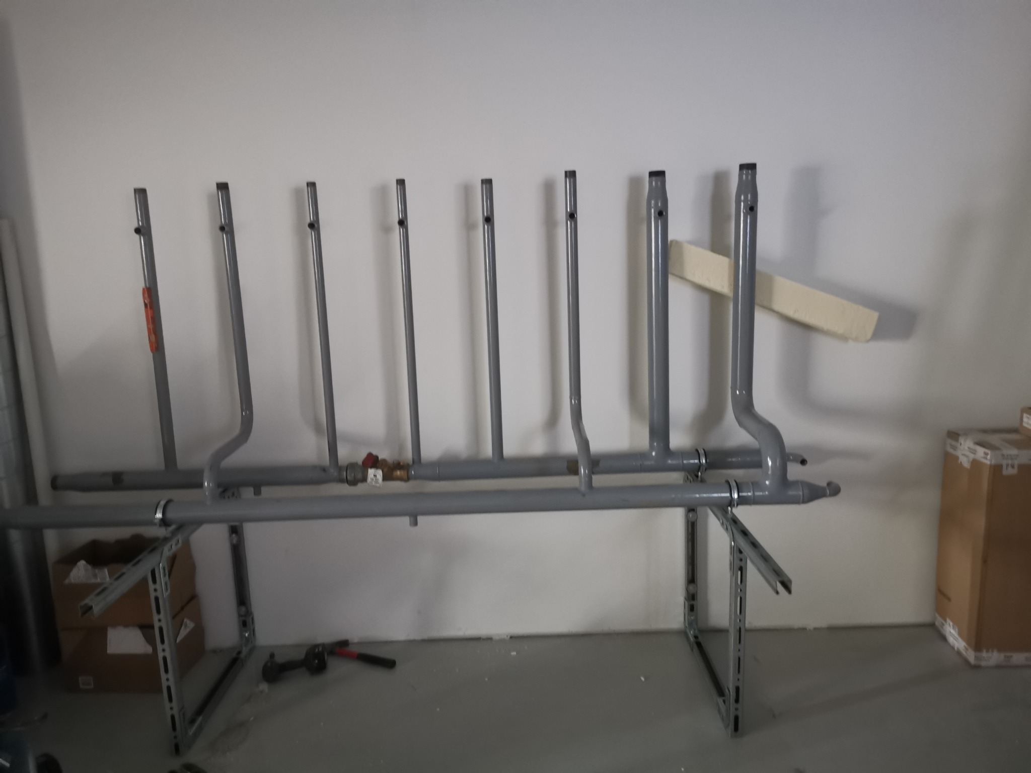 Szare rury instalacji grzewczej zamontowane na metalowych wspornikach, widoczny zawór i narzędzia na podłodze w surowym pomieszczeniu.