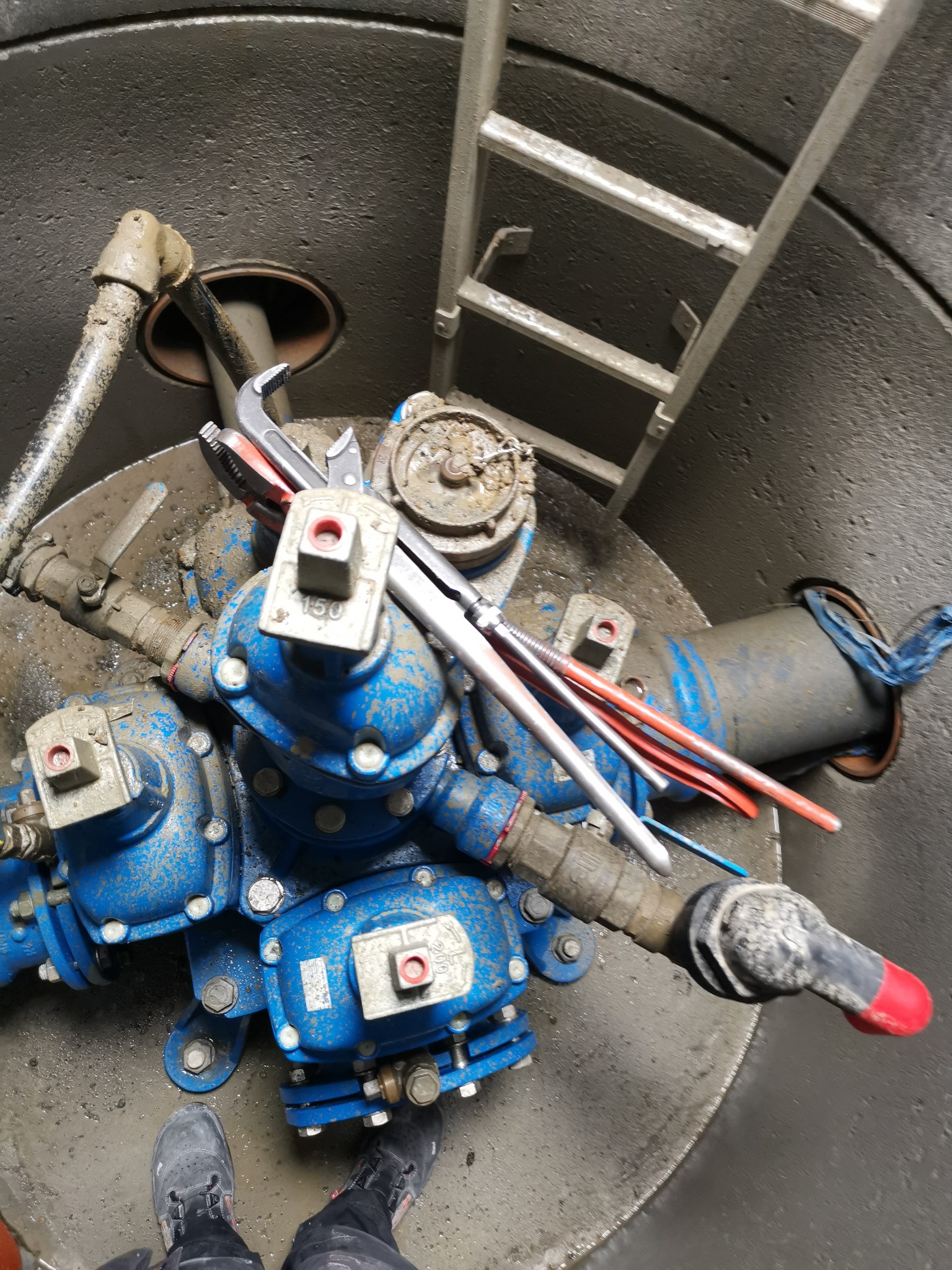 Wnętrze betonowej studzienki z niebieskimi zaworami wodnymi, rurami, kluczami hydraulicznymi i drabinką, widok z góry na instalację i obuwie hydraulika.