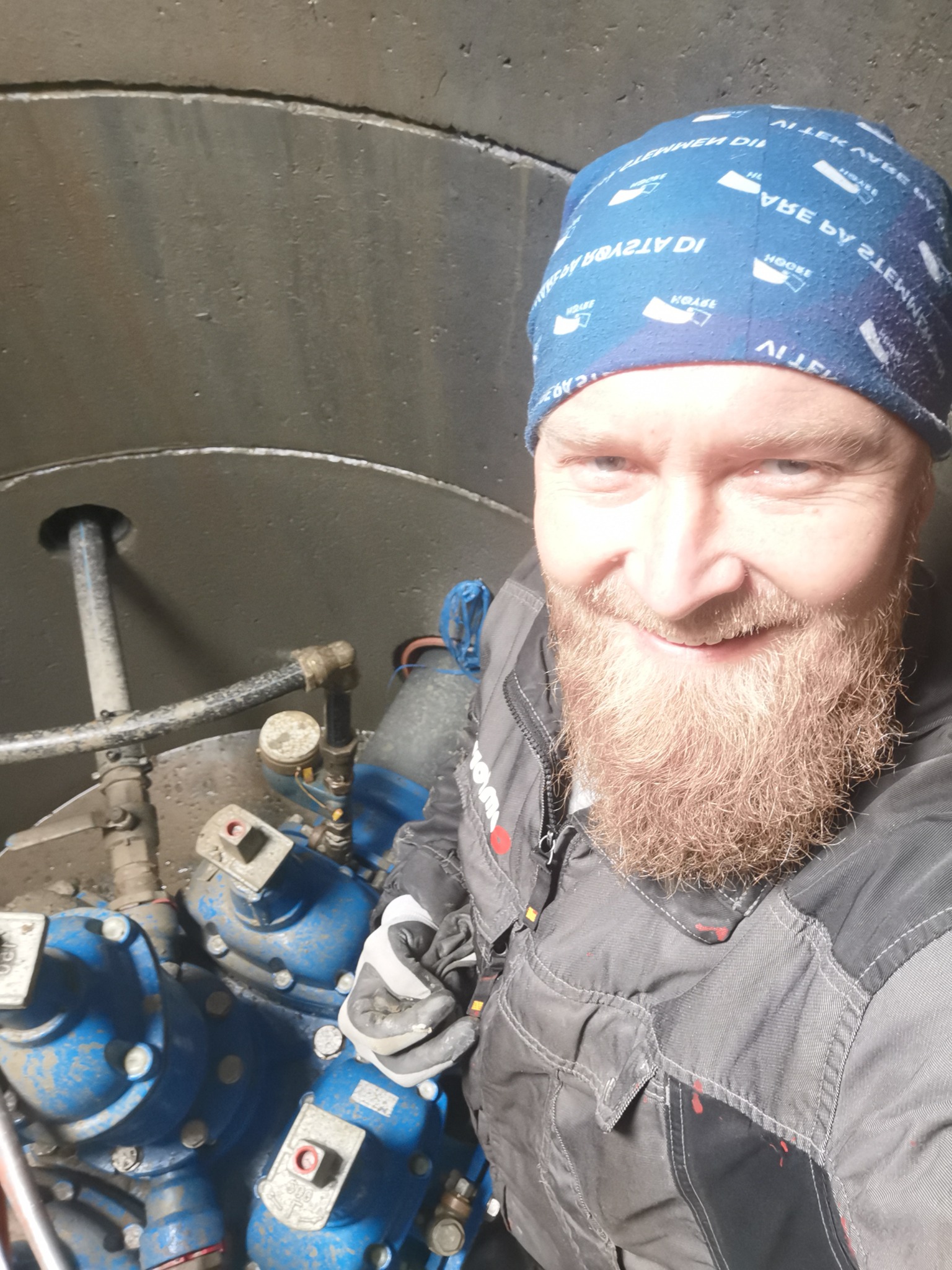 Uśmiechnięty hydraulik w studzience kanalizacyjnej pozuje do selfie na tle niebieskich zaworów i rur, ubrany w roboczy kombinezon i bandanę.