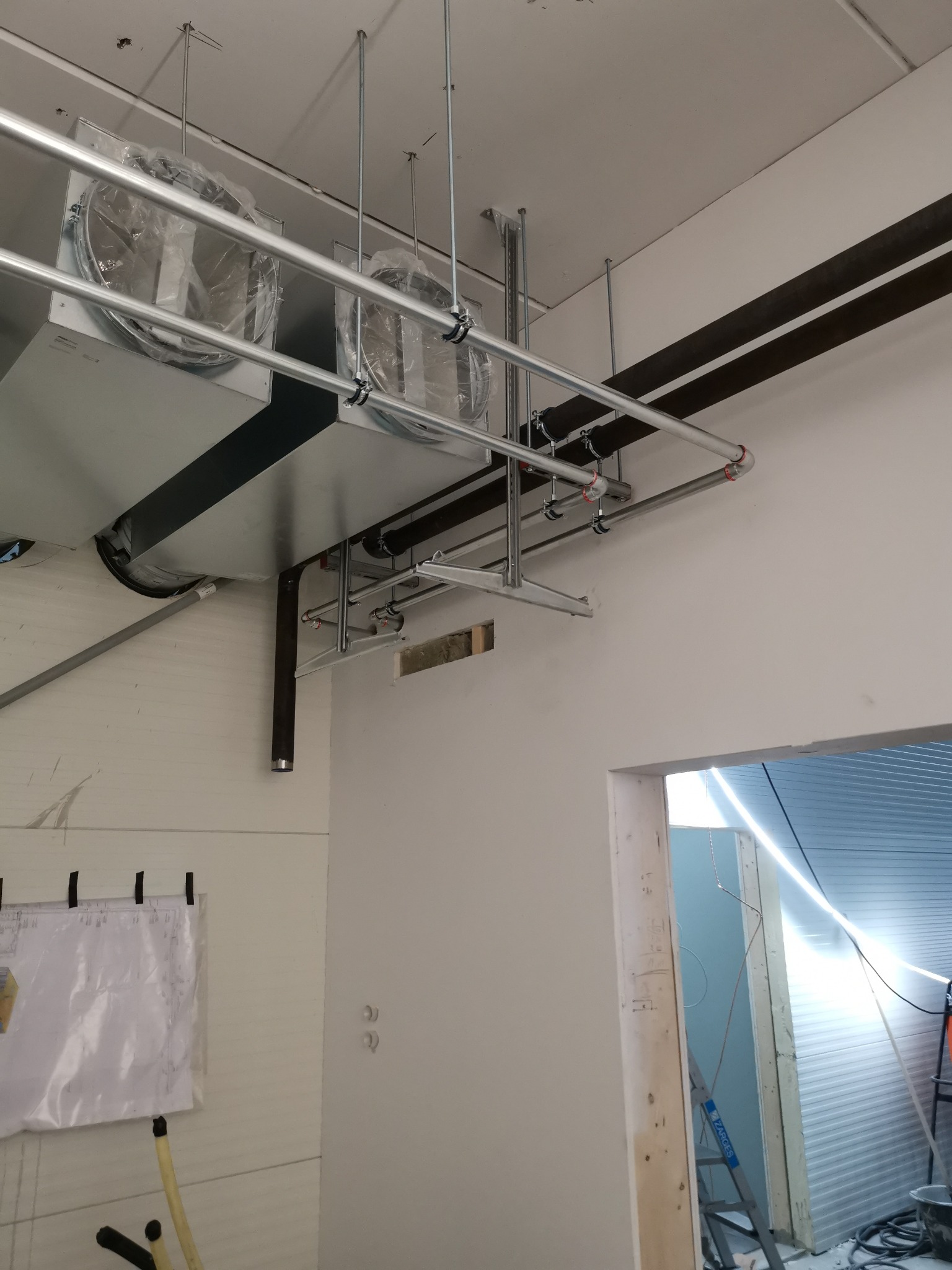 Widok na sufit z zamontowanymi elementami wentylacji, rurami i instalacjami podwieszonymi na metalowych prętach, częściowo jeszcze zabezpieczone folią ochronną, w tle widoczne niedokończone wnętrze...