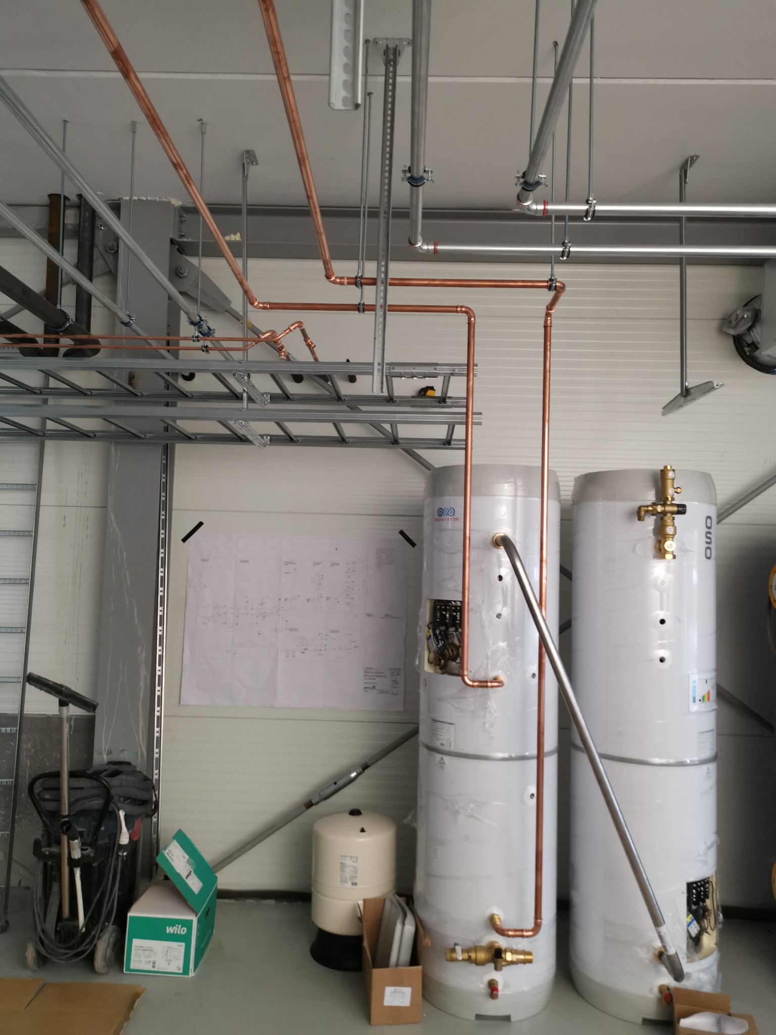 Dwa białe bojlery owinięte folią, połączone miedzianymi rurami, z widocznymi zaworami i elementami instalacji, na tle białej ściany i metalowej konstrukcji sufitowej.