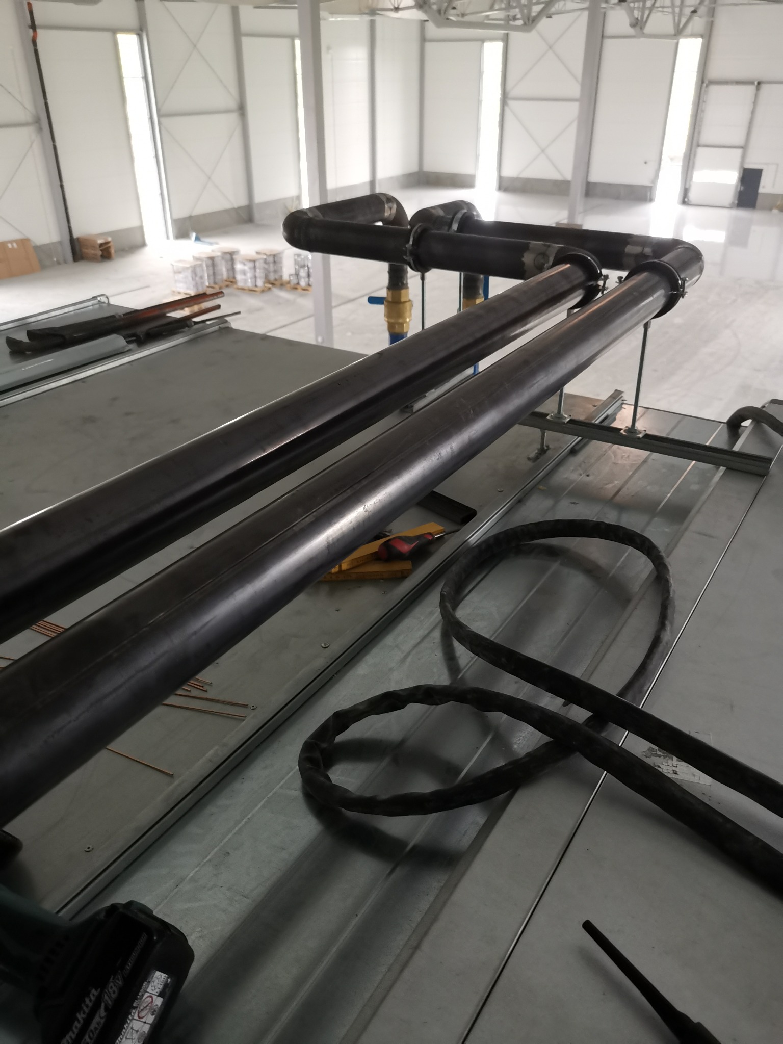 Spawane rury instalacji przemysłowej w hali, widoczne narzędzia i pozostałości po spawaniu, w tle duże okna doświetlające.