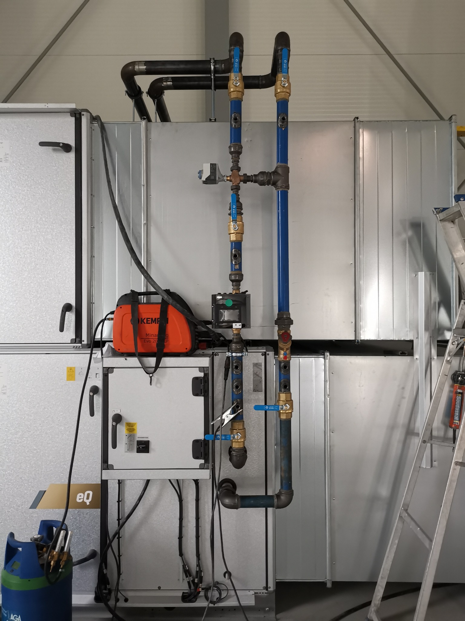 Instalacja przemysłowa z rurami w kolorze niebieskim i mosiężnym, zaworami, spawarką Kemppi Minaarc Evo 200 i butlą z gazem AGA, całość zamontowana na tle metalowej ściany z drabinką opartą o nią.