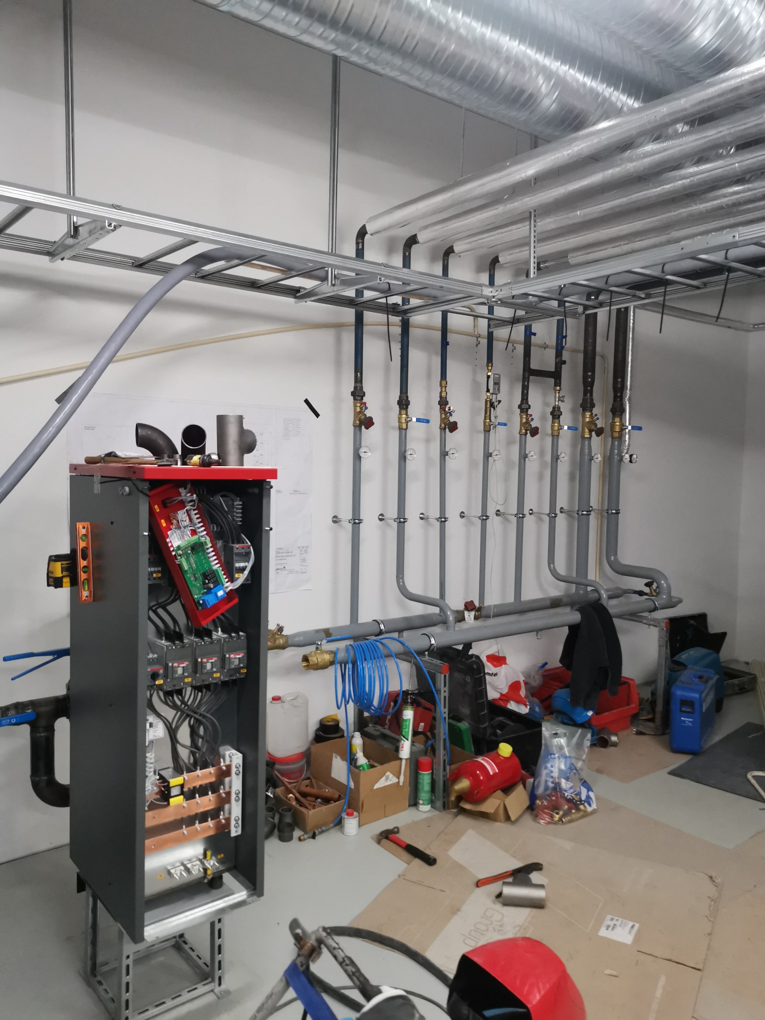 Otwarta szafa sterownicza z widoczną elektroniką i okablowaniem, na tle instalacji rur grzewczych i narzędzi na podłodze w pomieszczeniu technicznym.