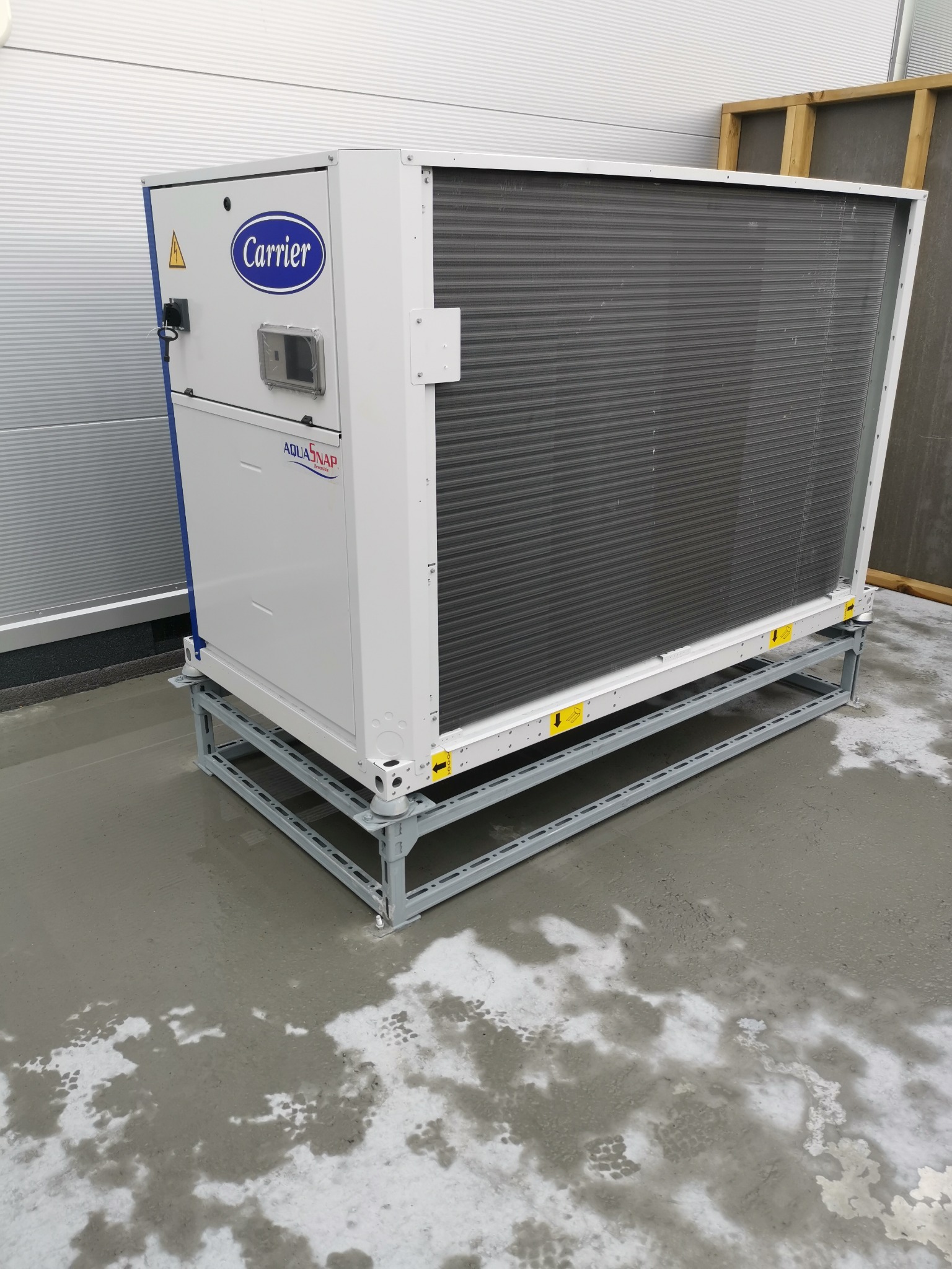 Klimatyzator przemysłowy Carrier AquaSnap na metalowej ramie, ustawiony na mokrej posadzce, z widocznym logo i tabliczką ostrzegawczą.