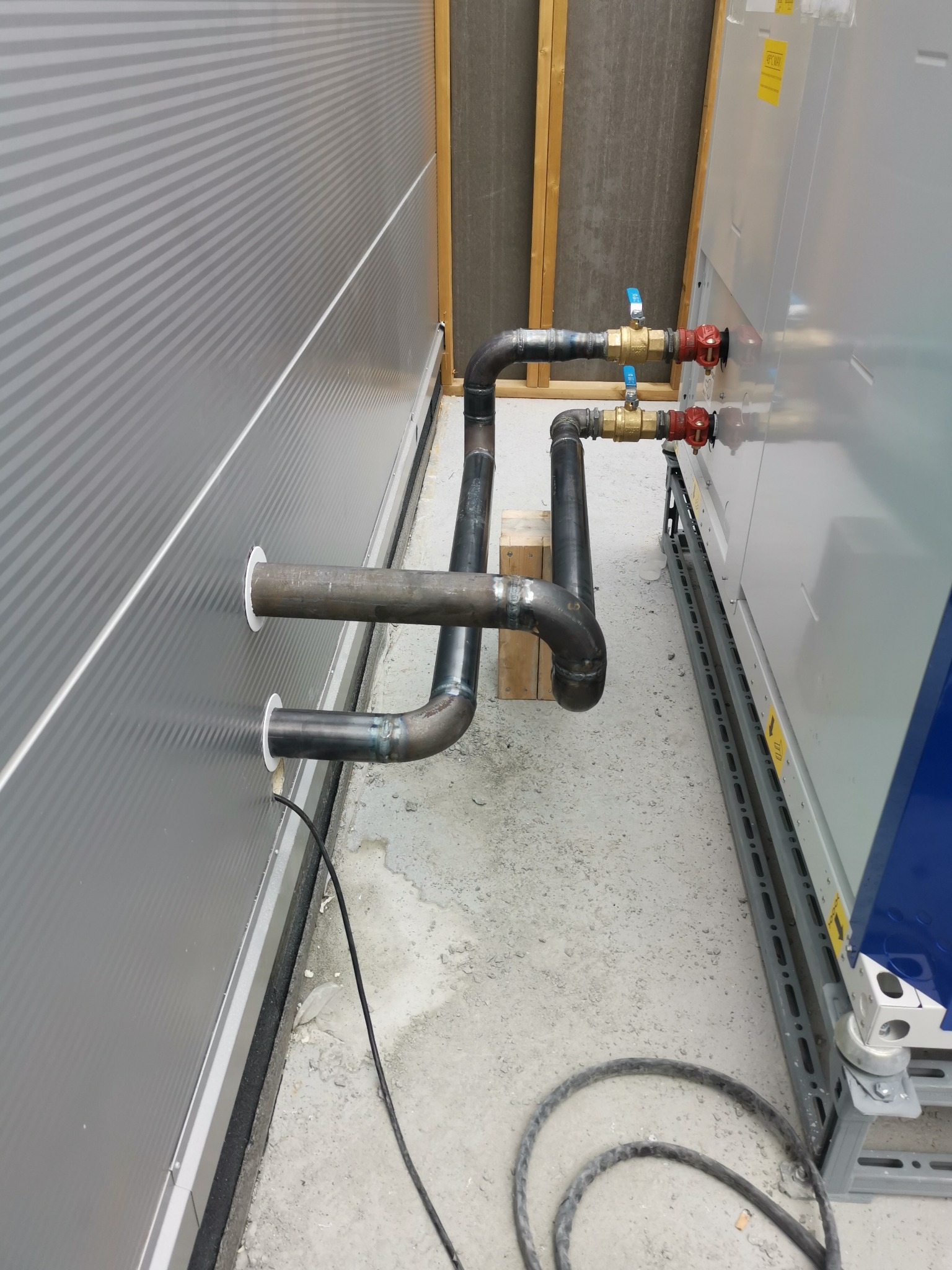 Zespawane rury instalacji grzewczej wchodzące przez otwory w panelowej ścianie zewnętrznej budynku, z zaworami odcinającymi na tle urządzenia grzewczego.