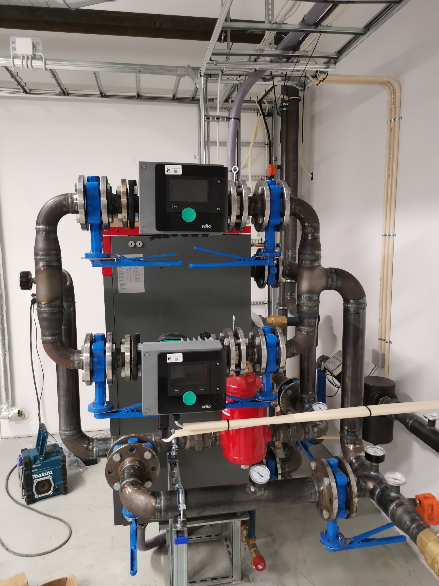 Kaskadowa instalacja grzewcza z dwoma piecami kondensacyjnymi Wilo, rurami stalowymi z niebieskimi zaworami kulowymi, manometrami i filtrem, zamontowana w schludnym pomieszczeniu technicznym...