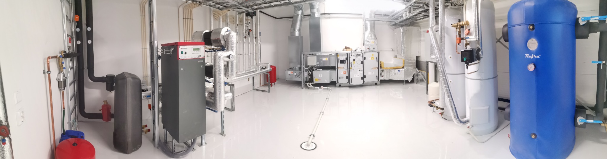 Szeroki kadr na kotłownię z instalacją grzewczą, wentylacyjną i zbiornikami wody, w jasnym pomieszczeniu z błyszczącą posadzką.