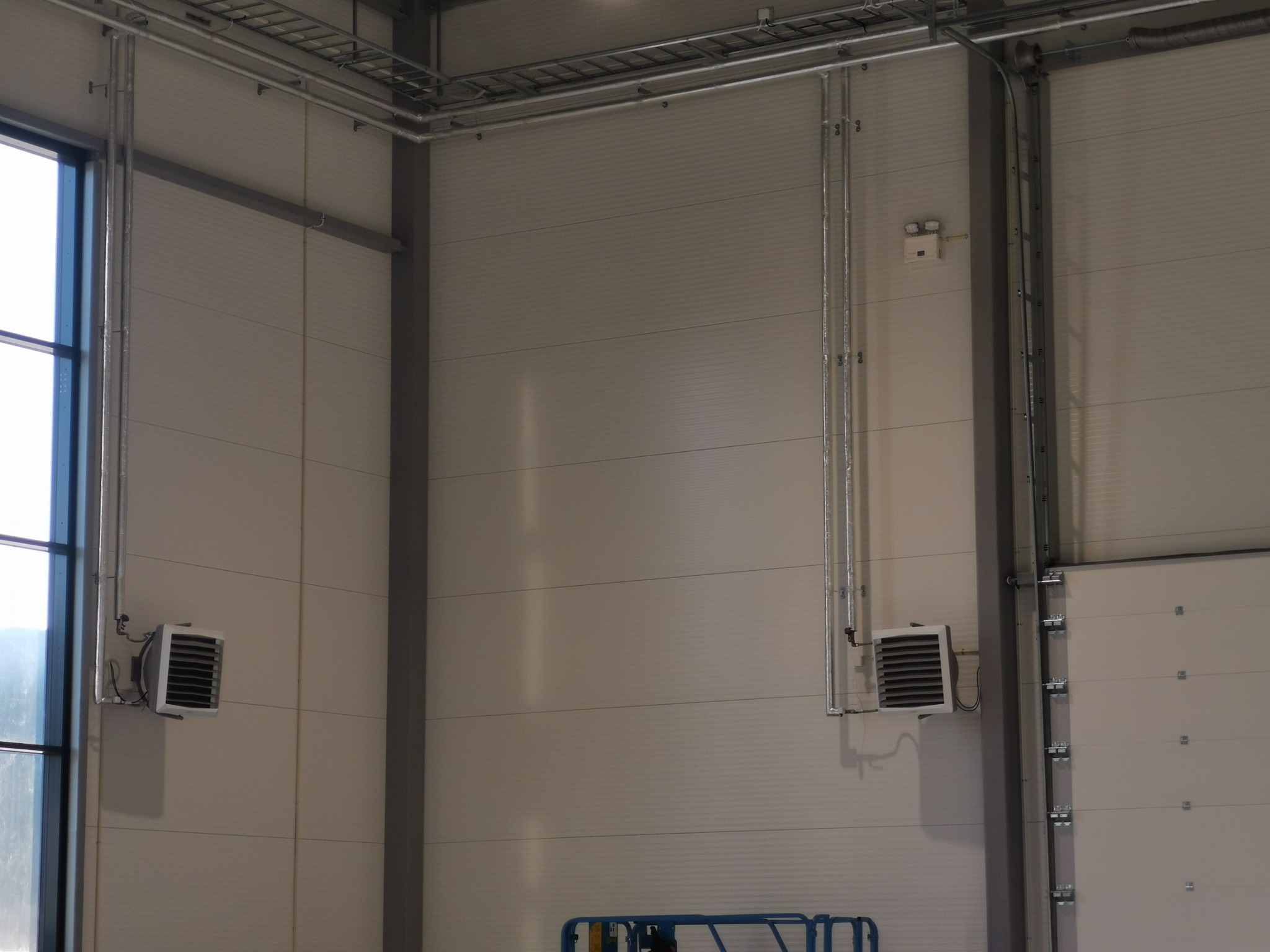 Dwa przemysłowe grzejniki elektryczne zamontowane na ścianie hali, z widocznymi rurami zasilającymi i metalowymi konstrukcjami pod sufitem.