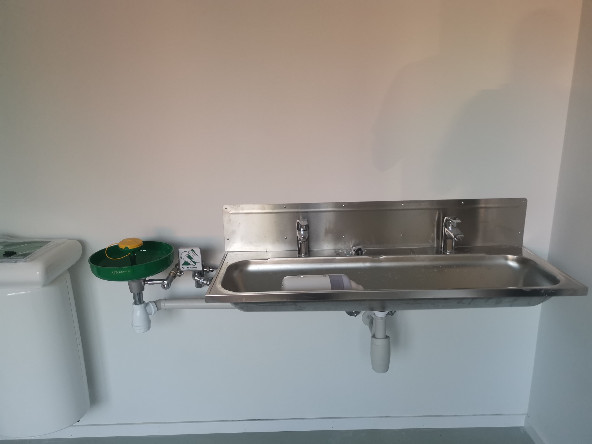 Zlew ze stali nierdzewnej z zamontowanym obok awaryjnym natryskiem do oczu, widoczne białe rury odpływowe i przymocowane do ściany w pomieszczeniu o jasnych ścianach.