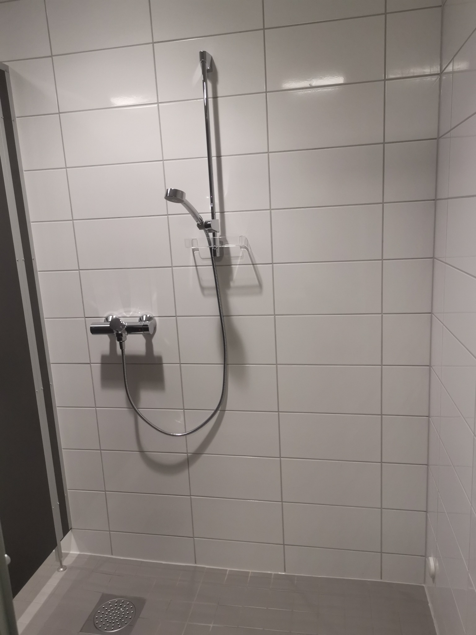 Nowoczesna kabina prysznicowa z białymi płytkami, chromowaną armaturą i odpływem liniowym w szarej posadzce.