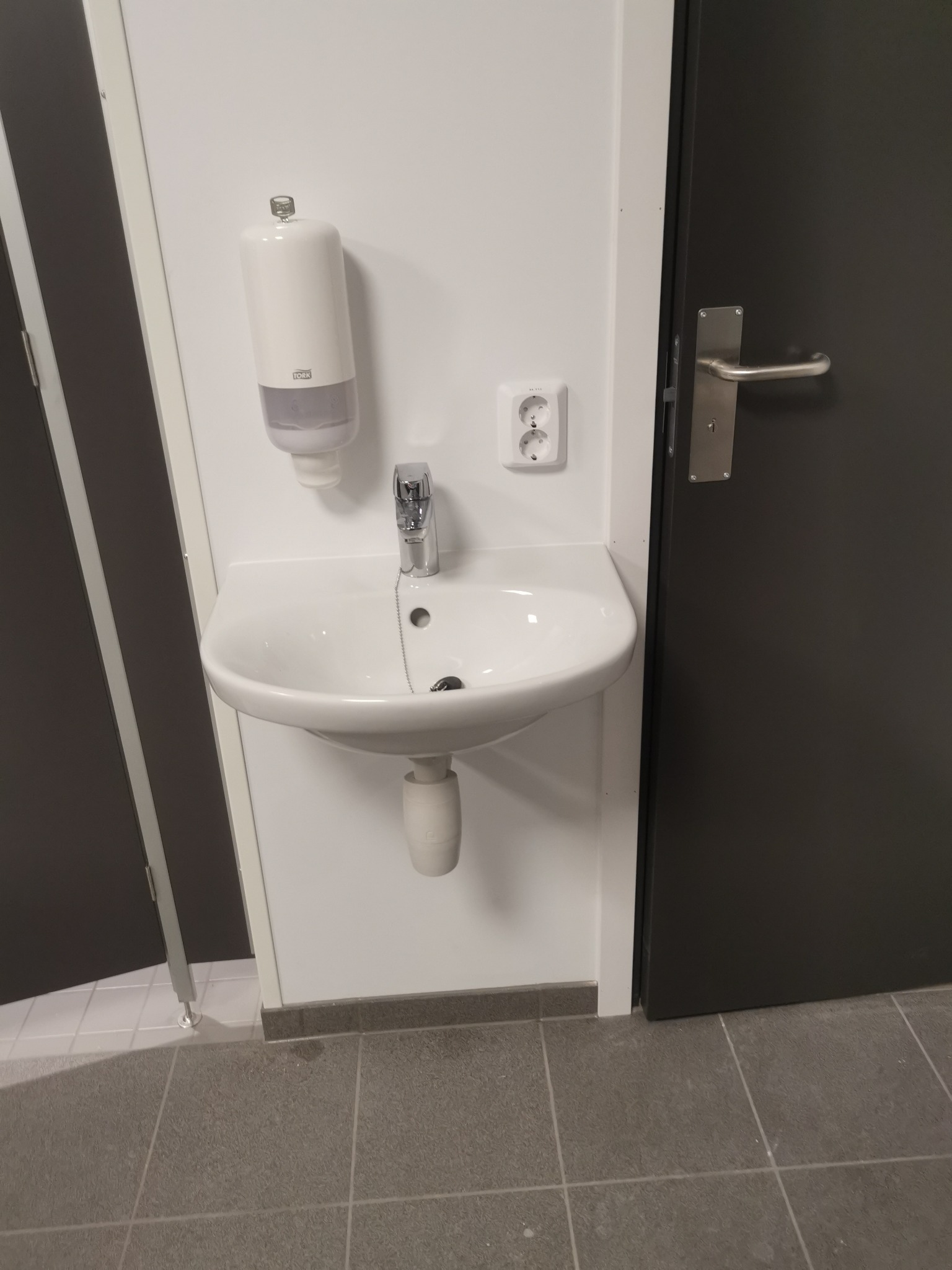 Biała umywalka zamontowana na ścianie w łazience, obok dozownik mydła i gniazdko elektryczne, widoczne fragmenty ciemnych drzwi i szarej podłogi.