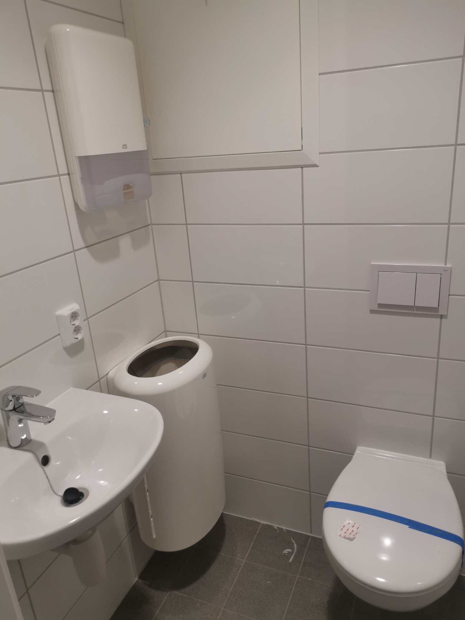 Nowoczesna, minimalistyczna łazienka w jasnych płytkach z umywalką, toaletą z niebieską taśmą zabezpieczającą i podtynkowym zbiornikiem, uzupełniona o podajnik na ręczniki papierowe i gniazdko...