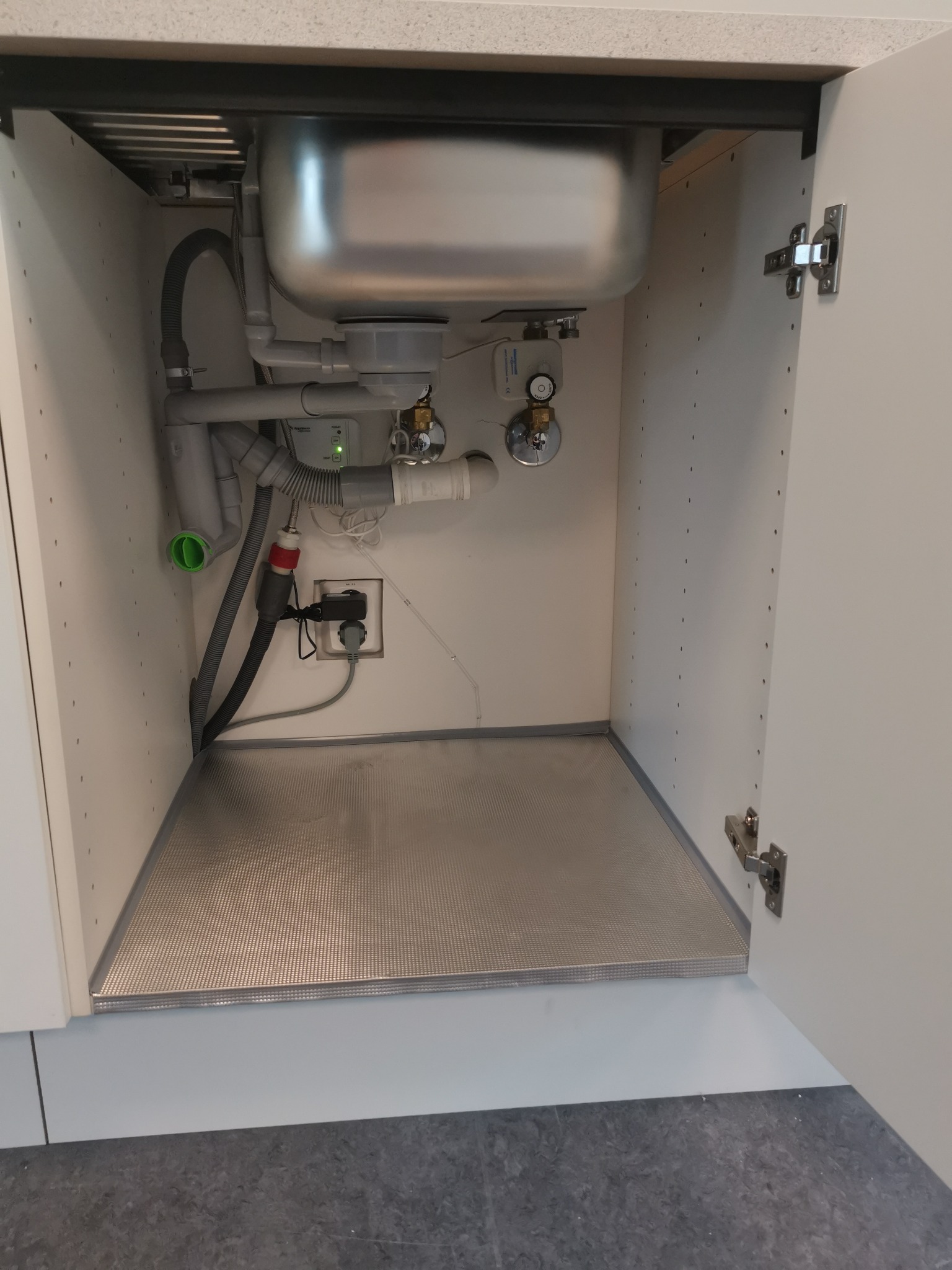 Instalacja odpływowa pod zlewem w szafce kuchennej z widocznymi rurami, syfonem, zaworami i matą zabezpieczającą dno szafki przed zalaniem.