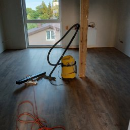 DomBud - Wnętrze pokoju z nową, ciemną podłogą drewnopodobną w trakcie sprzątania, widoczny żółty odkurzacz przemysłowy, listwa przypodłogowa w trakcie montażu oraz surowy, drewniany słup konstrukcyjny.