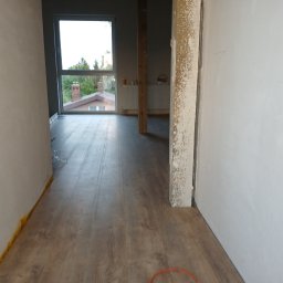 DomBud - Wnętrze w trakcie prac wykończeniowych: świeżo położona podłoga laminowana, surowe ściany przygotowane do malowania, widoczna izolacja przy podłodze, okno z widokiem na zieleń.