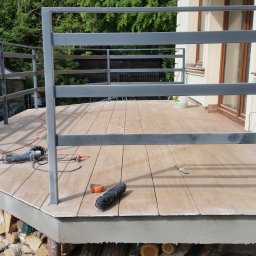 DomBud - Drewniany taras w trakcie budowy z szarą, metalową balustradą, widoczne narzędzia: szlifierka kątowa, miarka i pędzel, w tle dom i stos drewna.